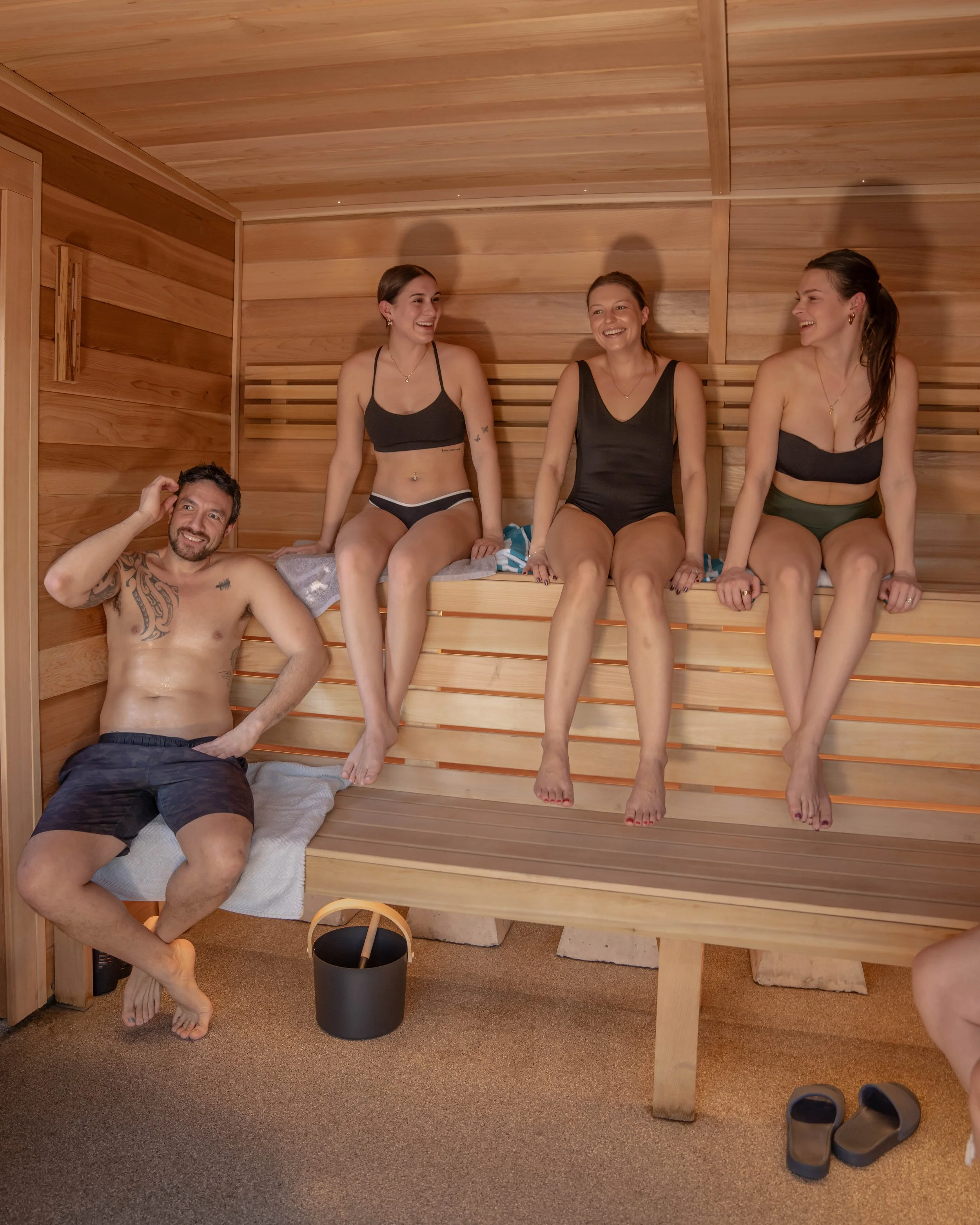 Four people relaxing in a wooden sauna with three women sitting on the top bench and one man sitting on the lower bench, all smiling and enjoying themselves.