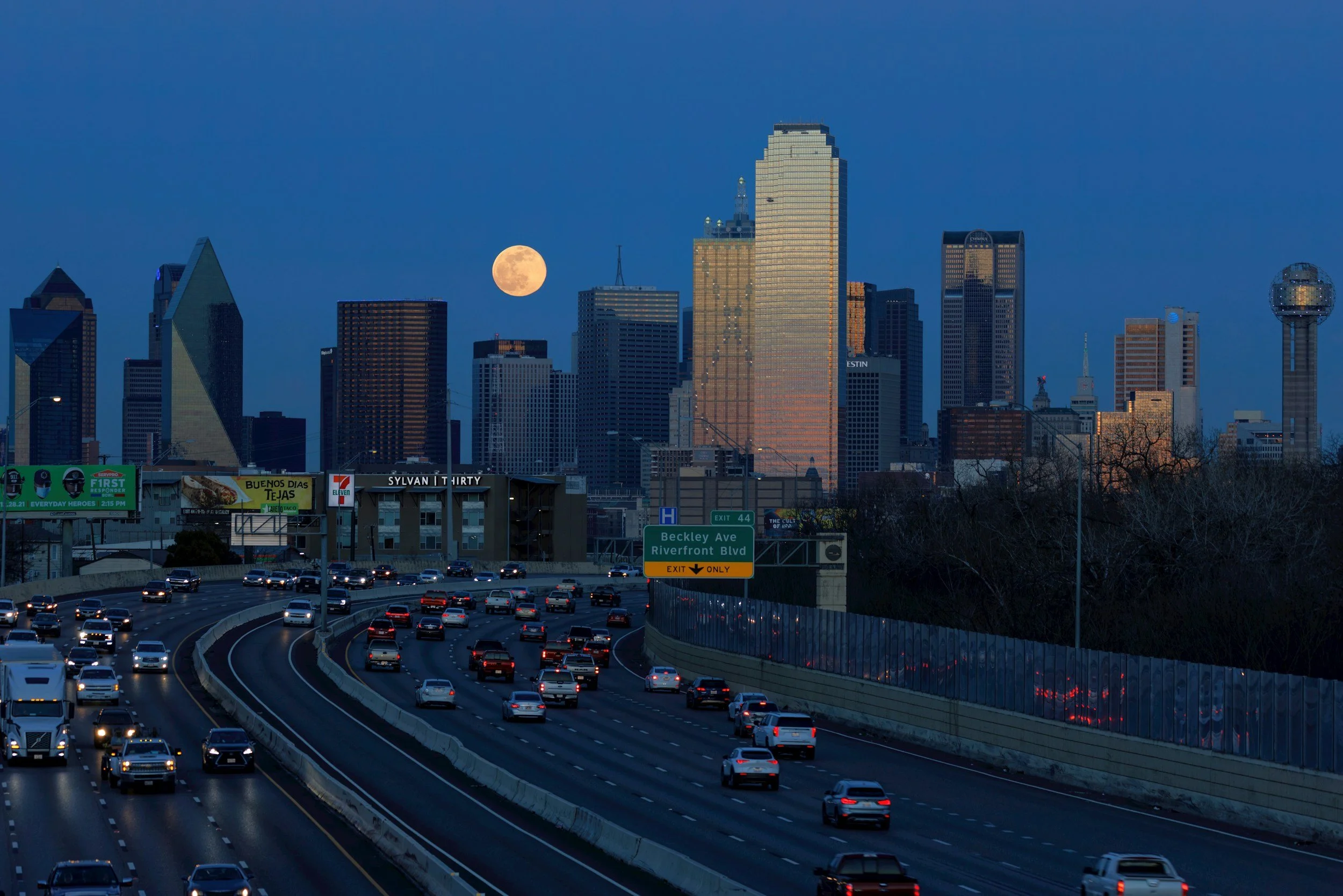 City skyline at dusk with the moon visible in the sky, traffic on a highway in the foreground, and tall skyscrapers in the background, including the Dallas skyline.