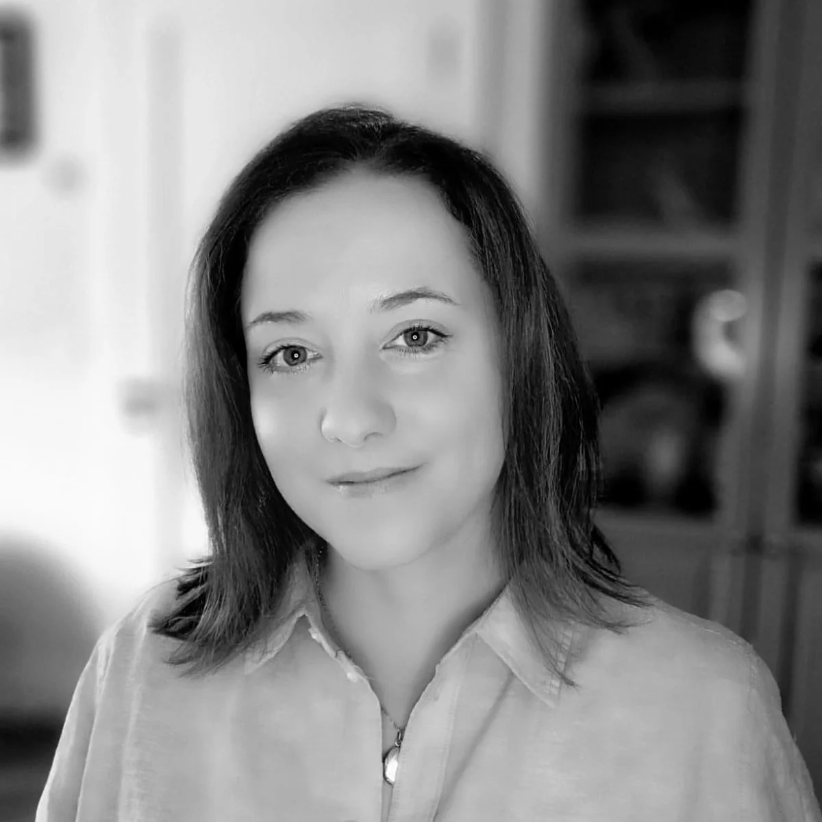 Black and white portrait of a woman with shoulder-length hair, wearing a light-colored shirt, indoors with a bookshelf in the background.