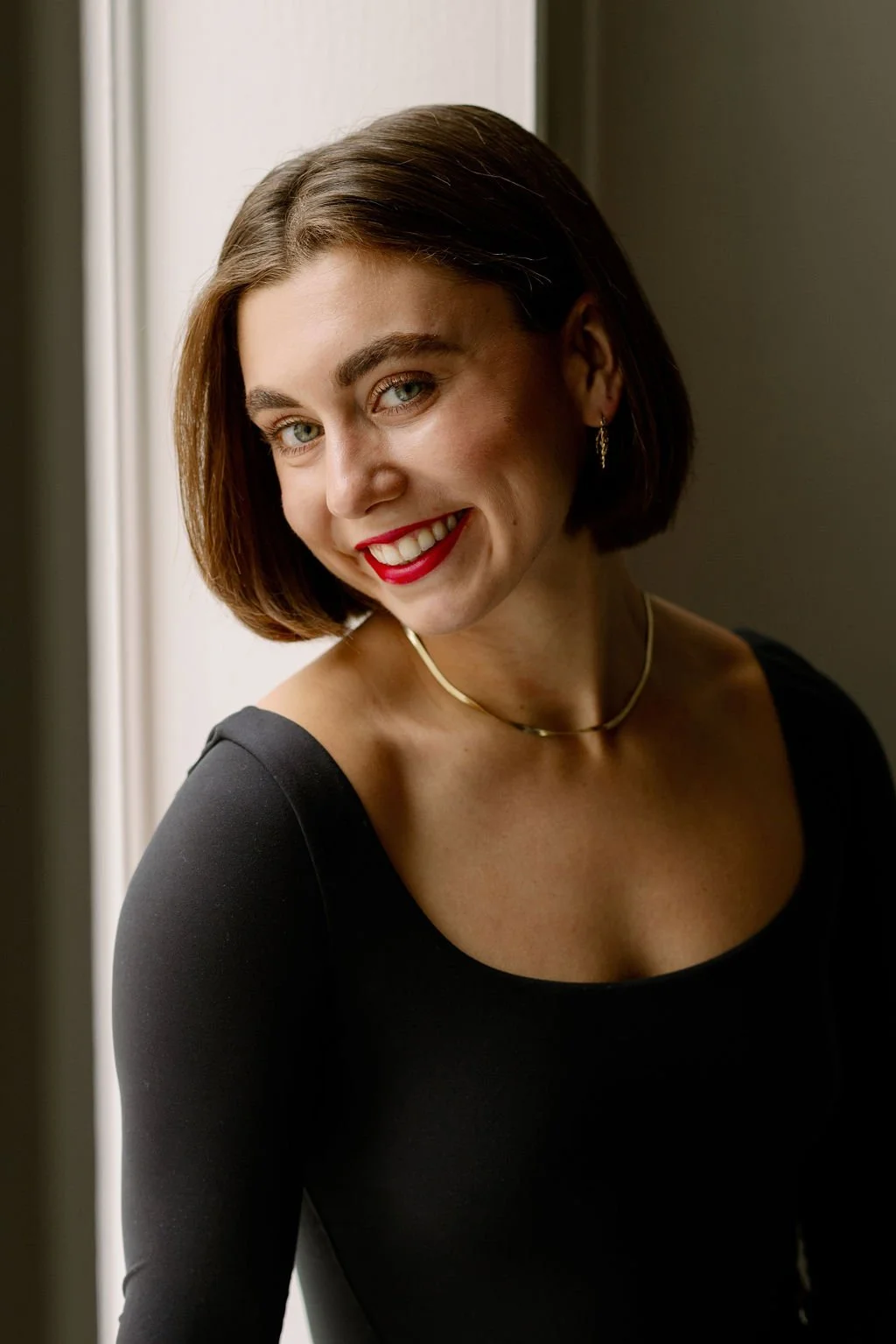 Close-up of a woman with short brown hair, green eyes, red lipstick, smiling, wearing a black top, gold hoop earrings, and a gold necklace, standing near a window with soft lighting.