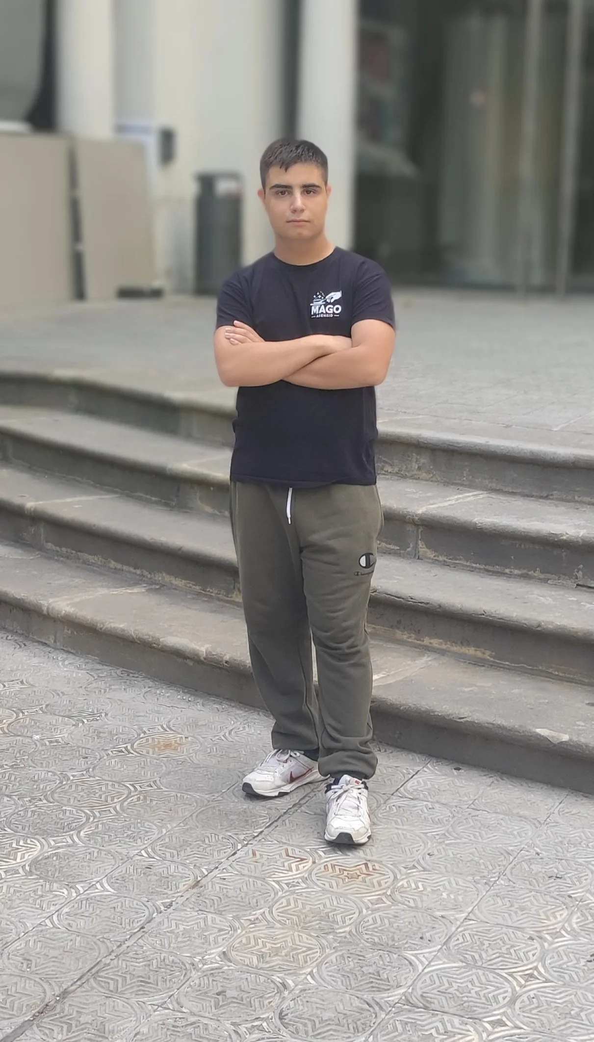 Joven de pie con brazos cruzados frente a escaleras en un entorno urbano, viste camiseta negra y pantalones deportivos grises.