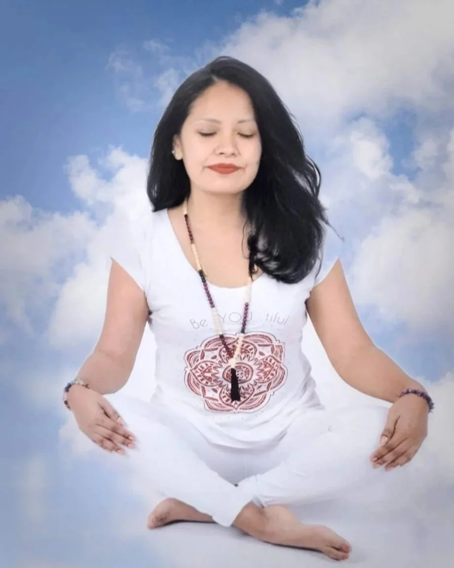 Mujer con cabello negro, ojos cerrados, en posición de meditación o yoga, con fondo de cielo con nubes, viste ropa blanca y usa accesorios de collar y pulseras.