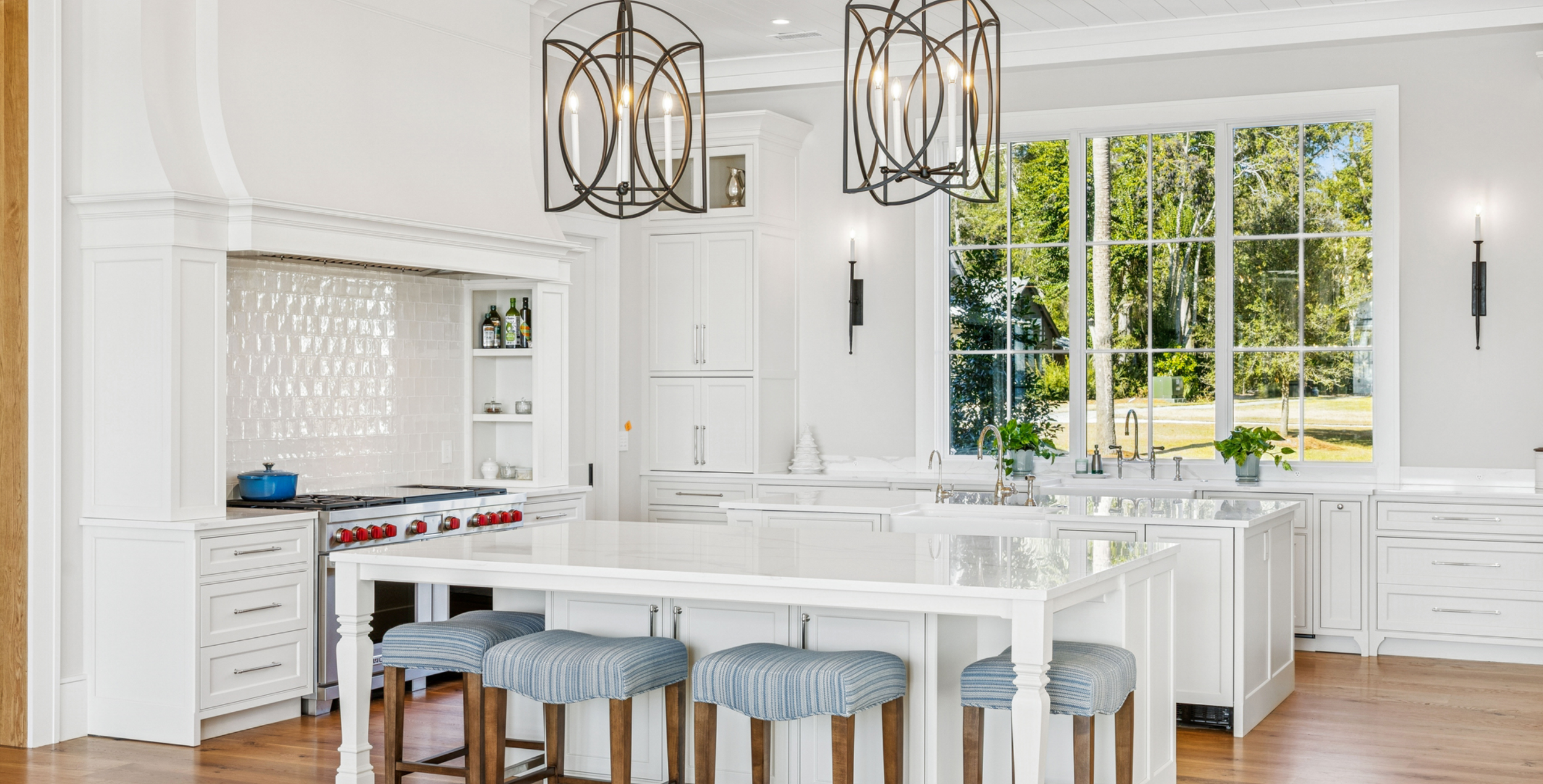 Modern white kitchen with large window, island, and Bosch oven, decorated with plants and pendant lighting.