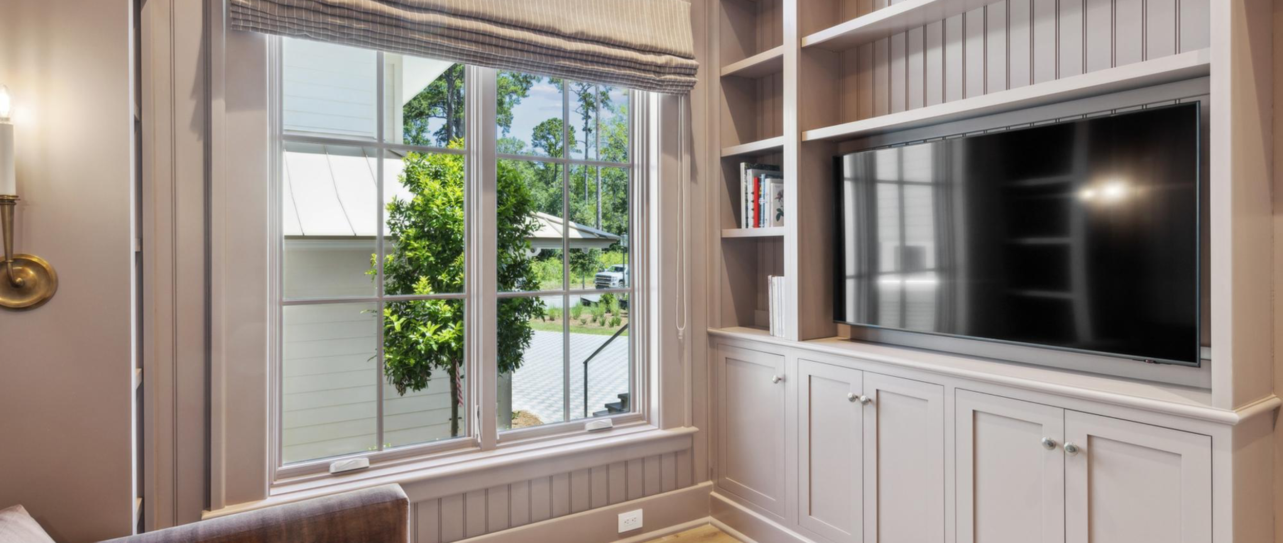 Living room corner with large window, view of green trees outside, built-in white wood cabinet with television on the right, bookshelves, and beige walls.