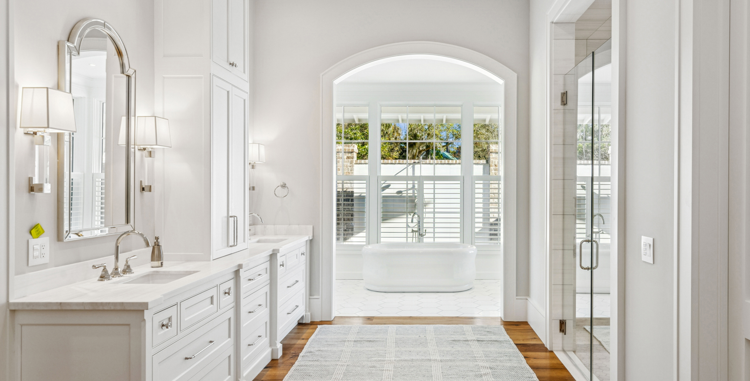 Bright white bathroom with double vanity sinks, mirrors, wall sconces, a bathtub by a large window, and a glass shower enclosure.