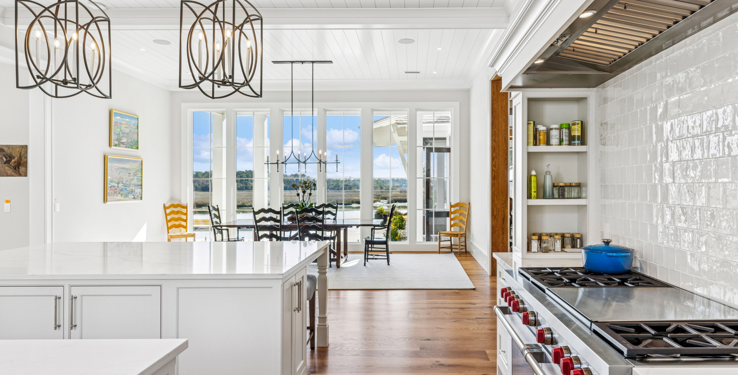 Bright kitchen with white cabinets, stainless steel stove, white tiled backsplash, and large windows overlooking a body of water with a blue sky and clouds.