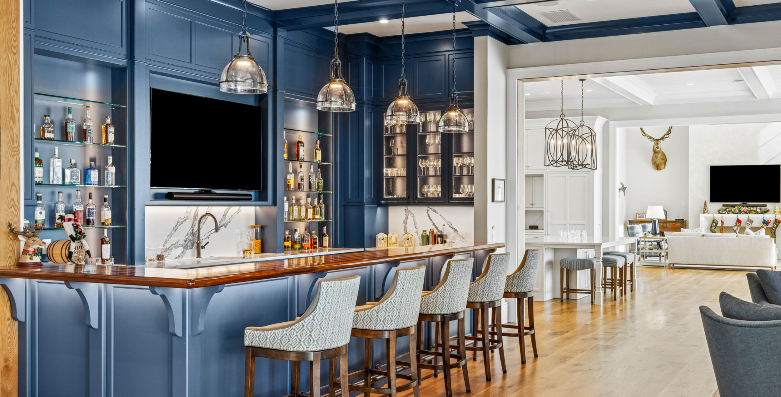 Interior of a modern living space with a blue kitchen bar area featuring a wooden countertop, bar stools with patterned upholstery, a large flat-screen TV, and a white area with a mounted deer head and holiday decorations.