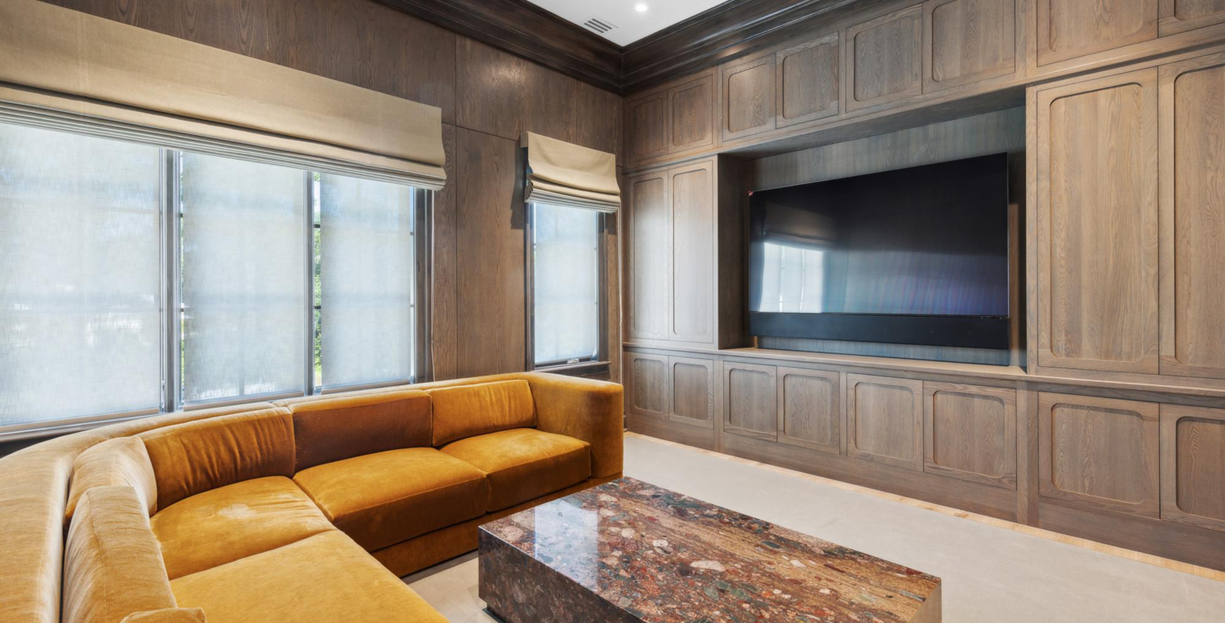 Living room with large windows covered by beige blinds, a mustard-colored sectional sofa, a coffee table with a multicolored stone top, and a large flat-screen TV mounted on a built-in wooden wall unit.