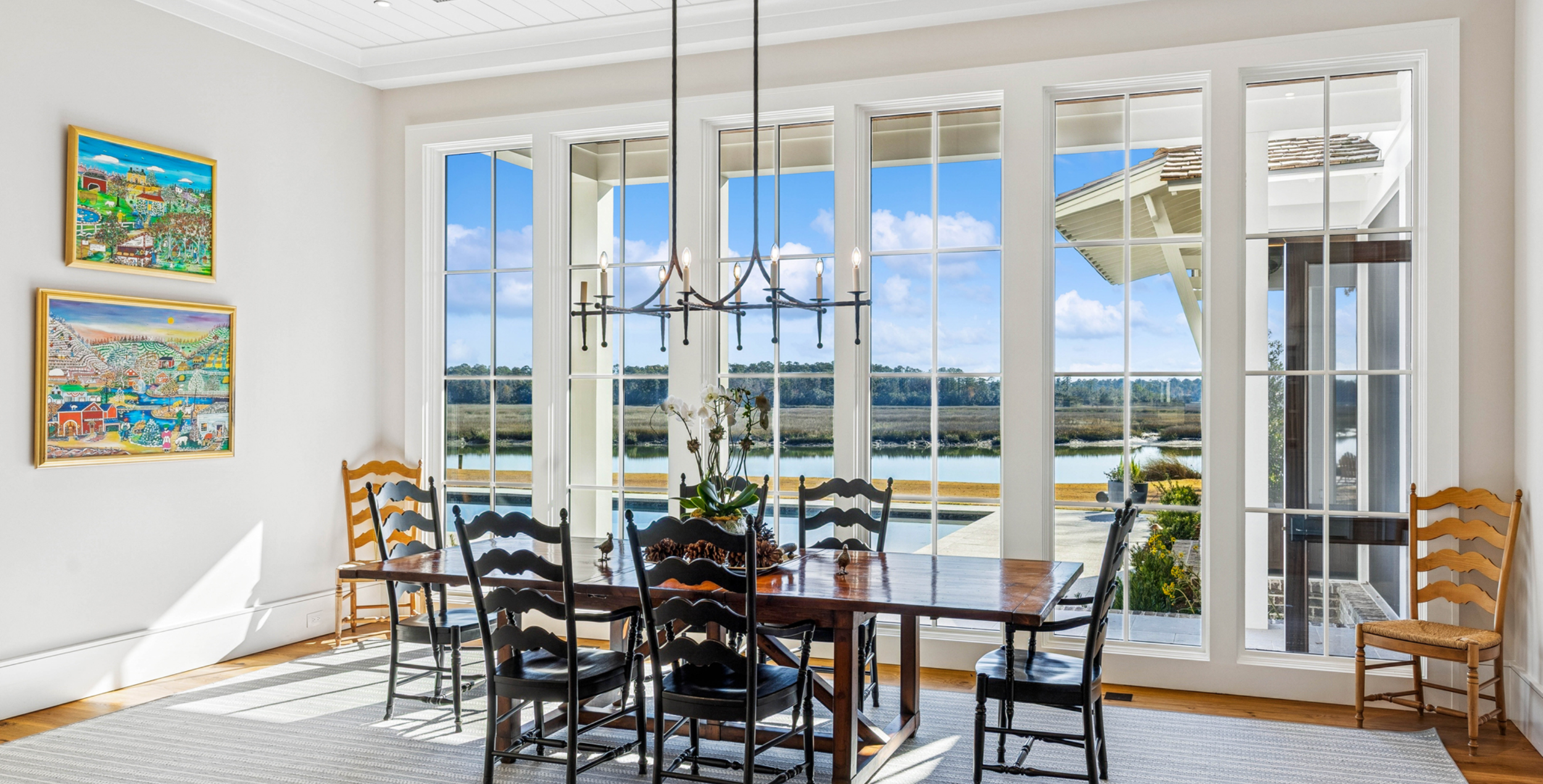 Dining room with a wooden table and black chairs, large windows showing a view of water and trees, artwork on the wall, and a black chandelier hanging above the table.