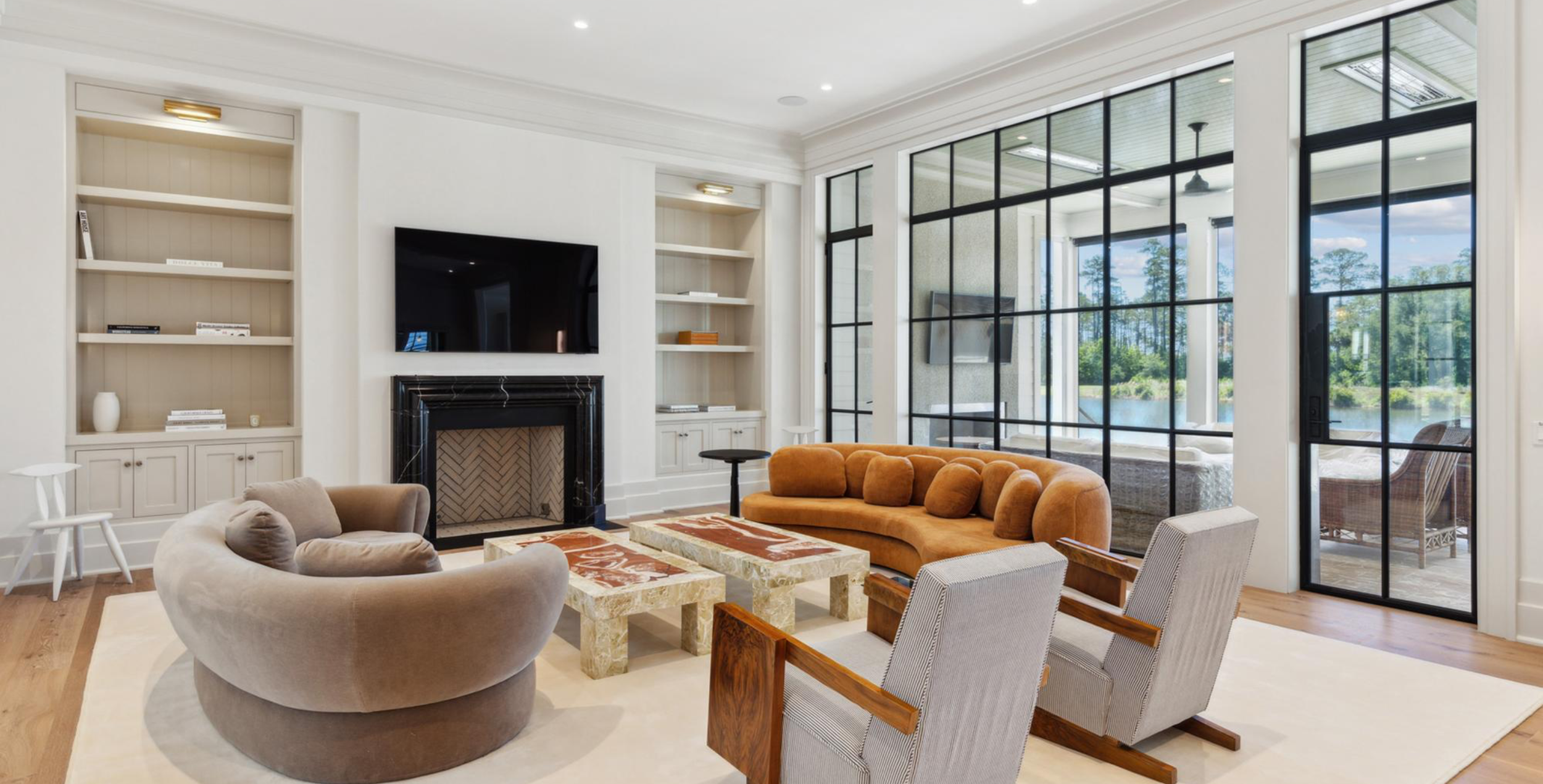 Modern living room with large windows, a fireplace, a flat screen TV, built-in shelves, and contemporary furniture including a curved beige sofa and wooden chairs.