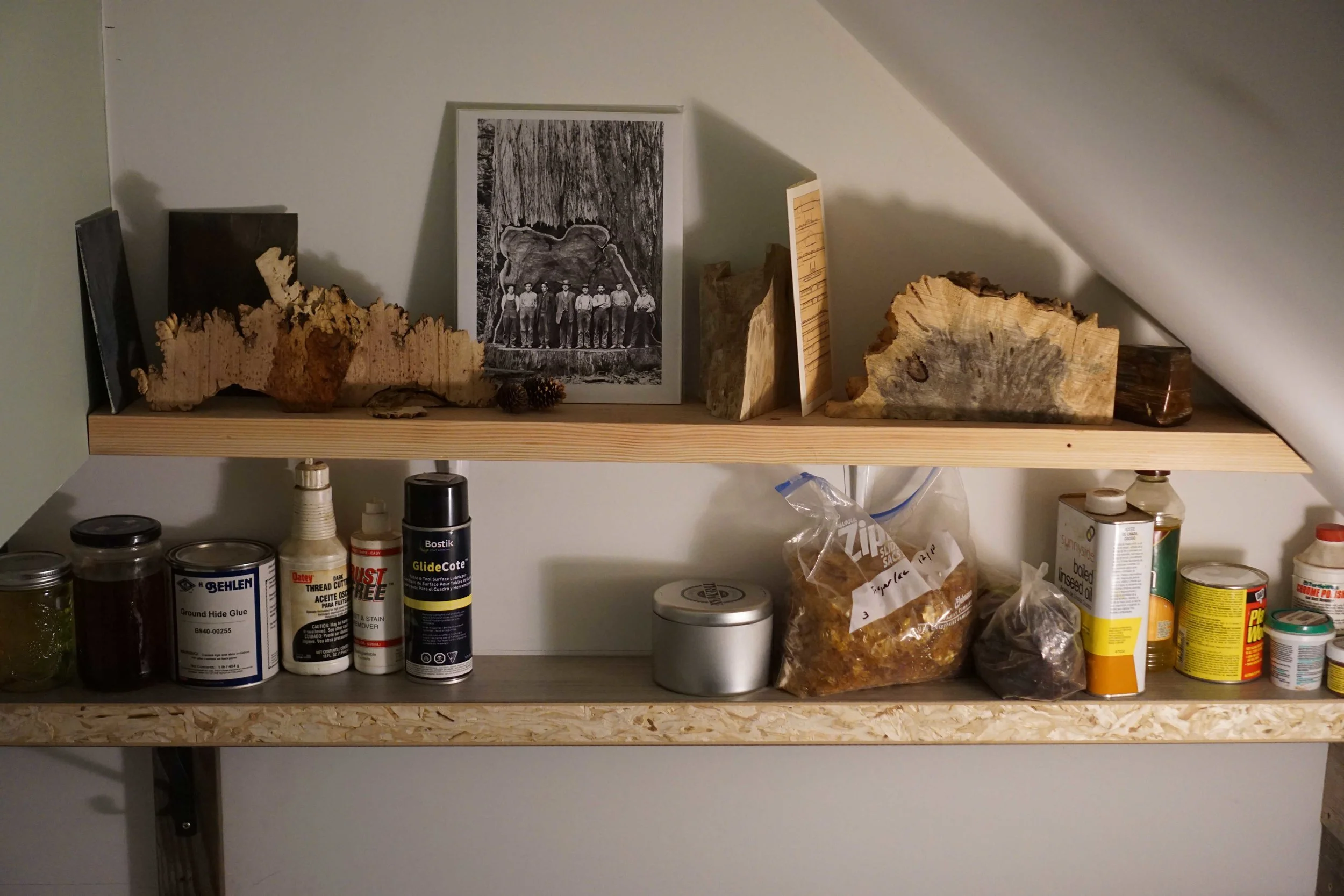 Two shelves with burl wood pieces, photo of lumberjacks, and guitar making supples.