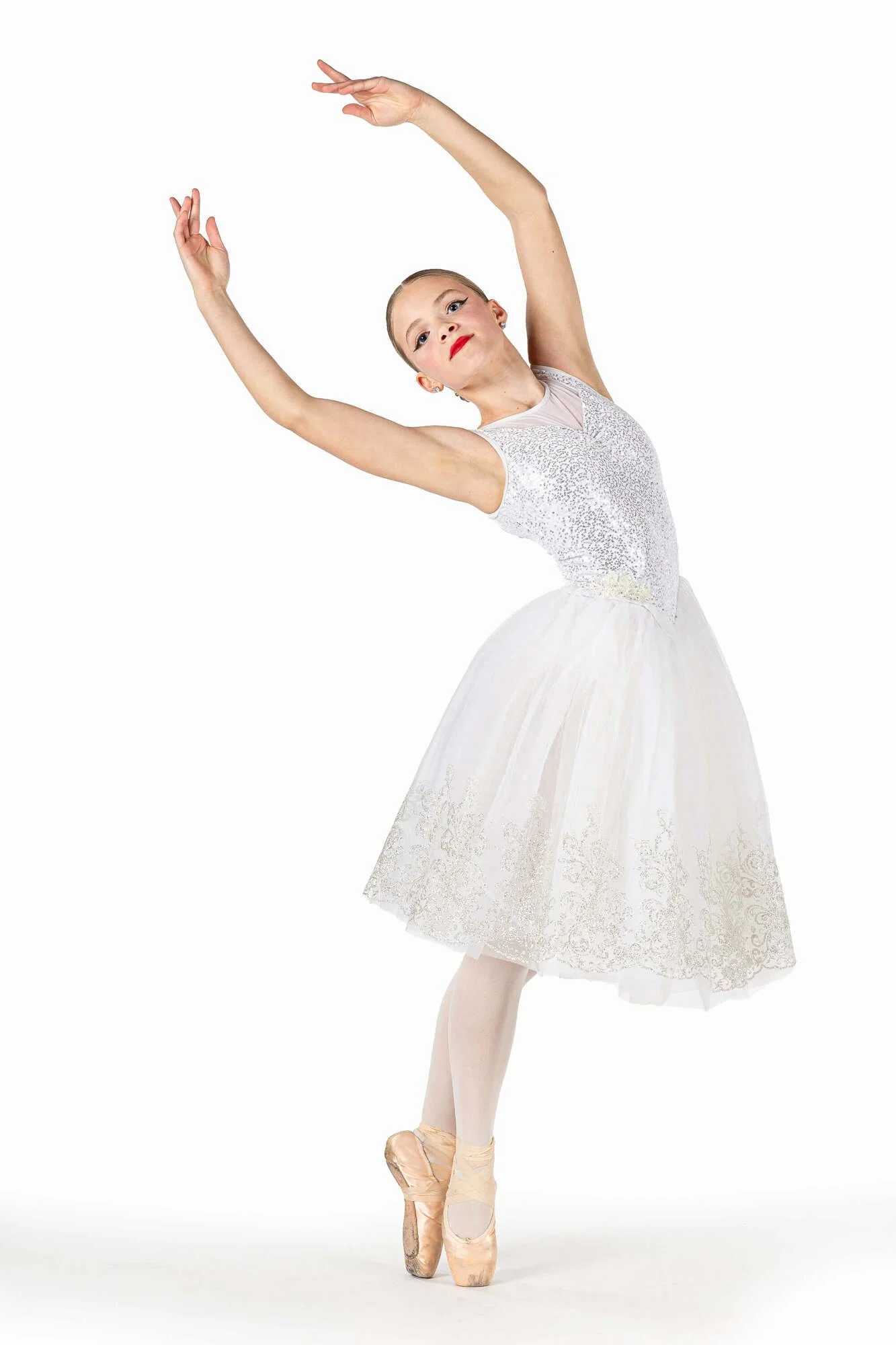 A young female ballet dancer in a white tutu and pointe shoes performing a ballet pose with her arms curved above her head against a white background.
