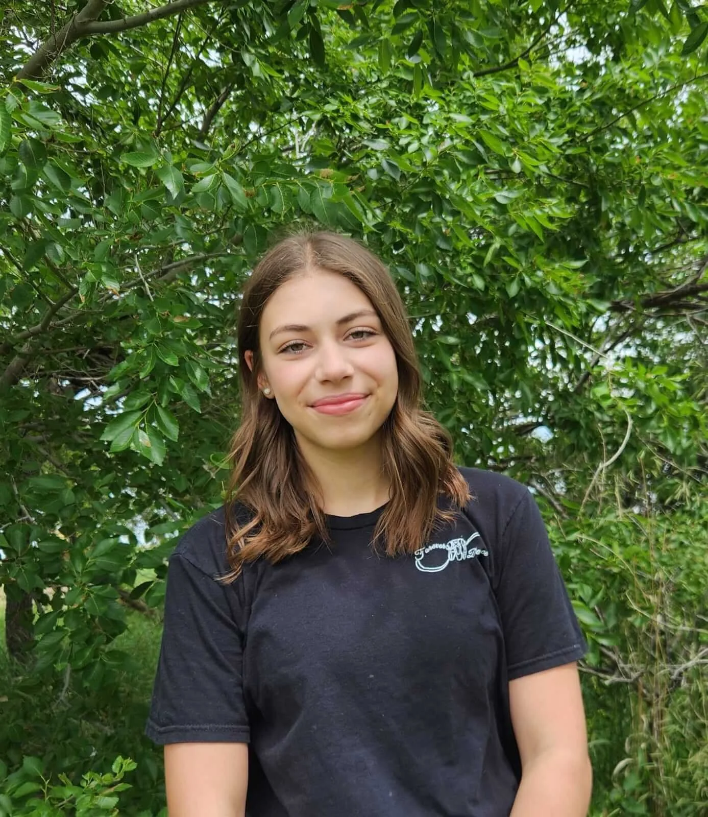 A young woman with brown hair wearing a black t-shirt standing outdoors in front of lush green foliage.