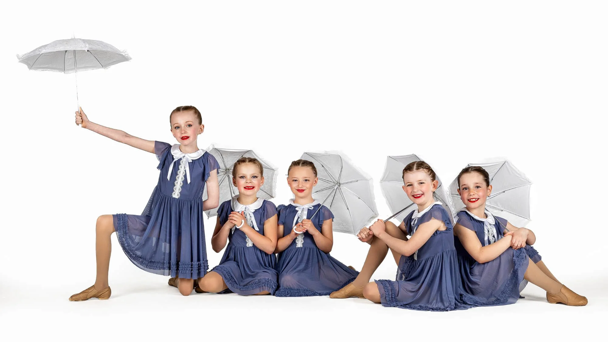 Group of five young girls dressed in matching blue dresses, sitting and kneeling on the floor, holding white umbrellas, posing for a photo in a studio with a white background.