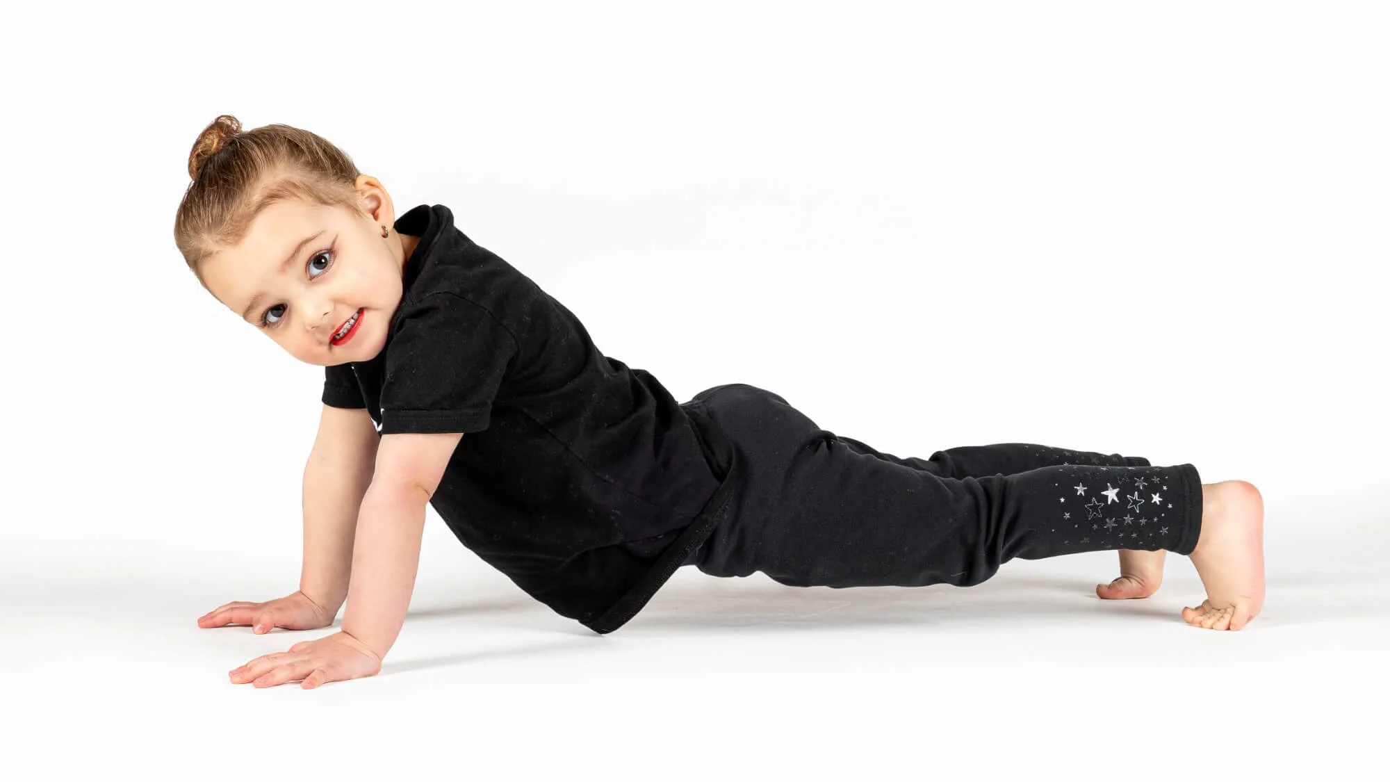 A young girl with light brown hair in a bun, wearing black clothing with star designs on her pants, performing a plank exercise on a white background, looking at the camera with a smile.