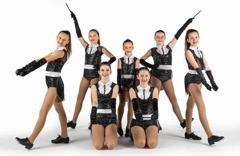 Group of seven young female dancers in black and white costumes with gloves, posing in the Forever Dance studio with a white background.