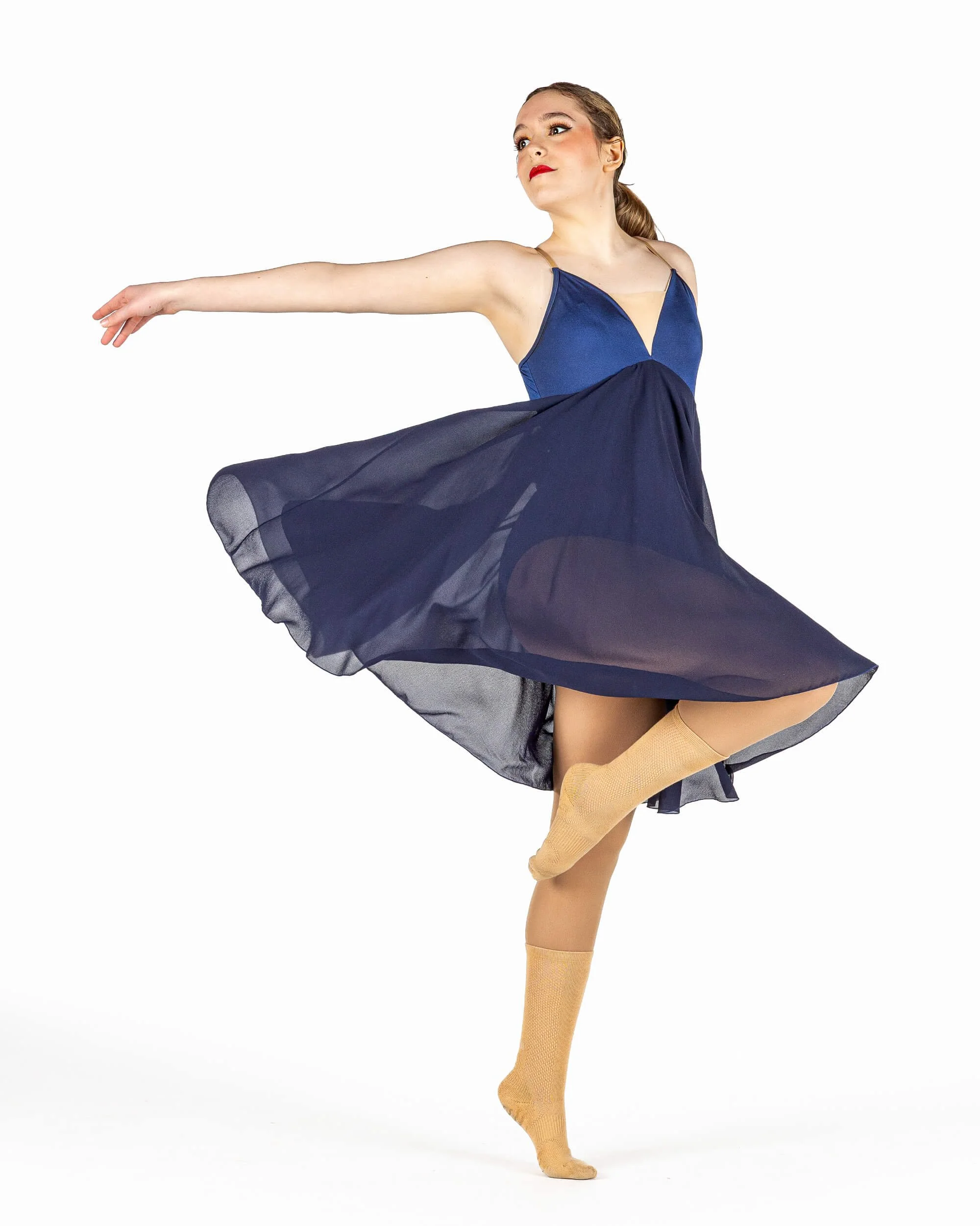 A ballet dancer performing a pose in a blue dress with a flowing skirt and beige socks, against a white background.