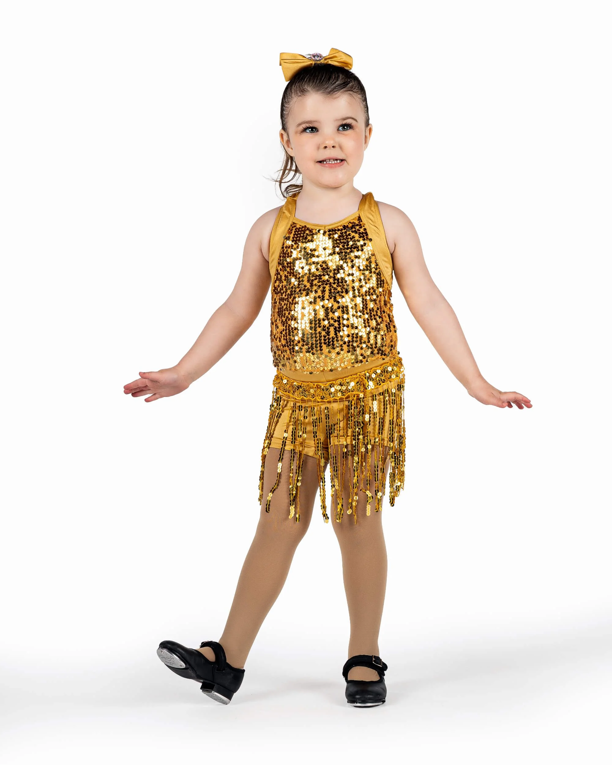 Young girl in a gold sequined and fringed dance costume, balancing on one leg with arms extended, against a white background.