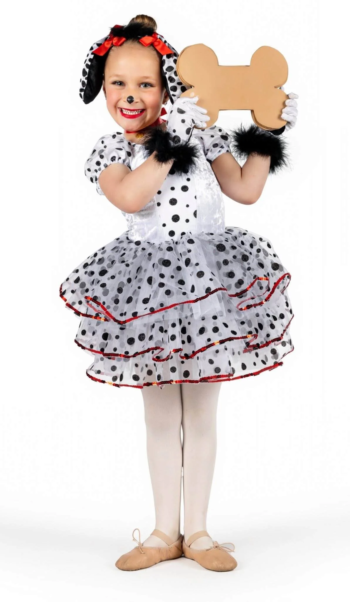 A young girl dressed in a Dalmatian puppy costume, holding a bone-shaped prop, smiling, with painted nose and dog-themed accessories.