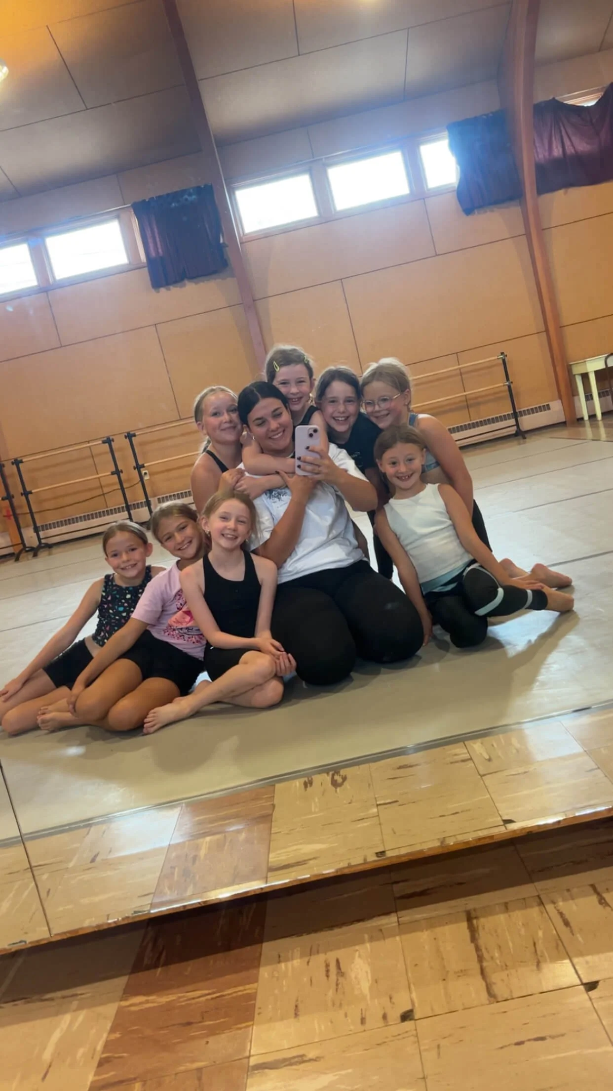 A group of young girls and a woman taking a selfie in the Forever Dance studio, all smiling and sitting on the floor in front of a mirror.