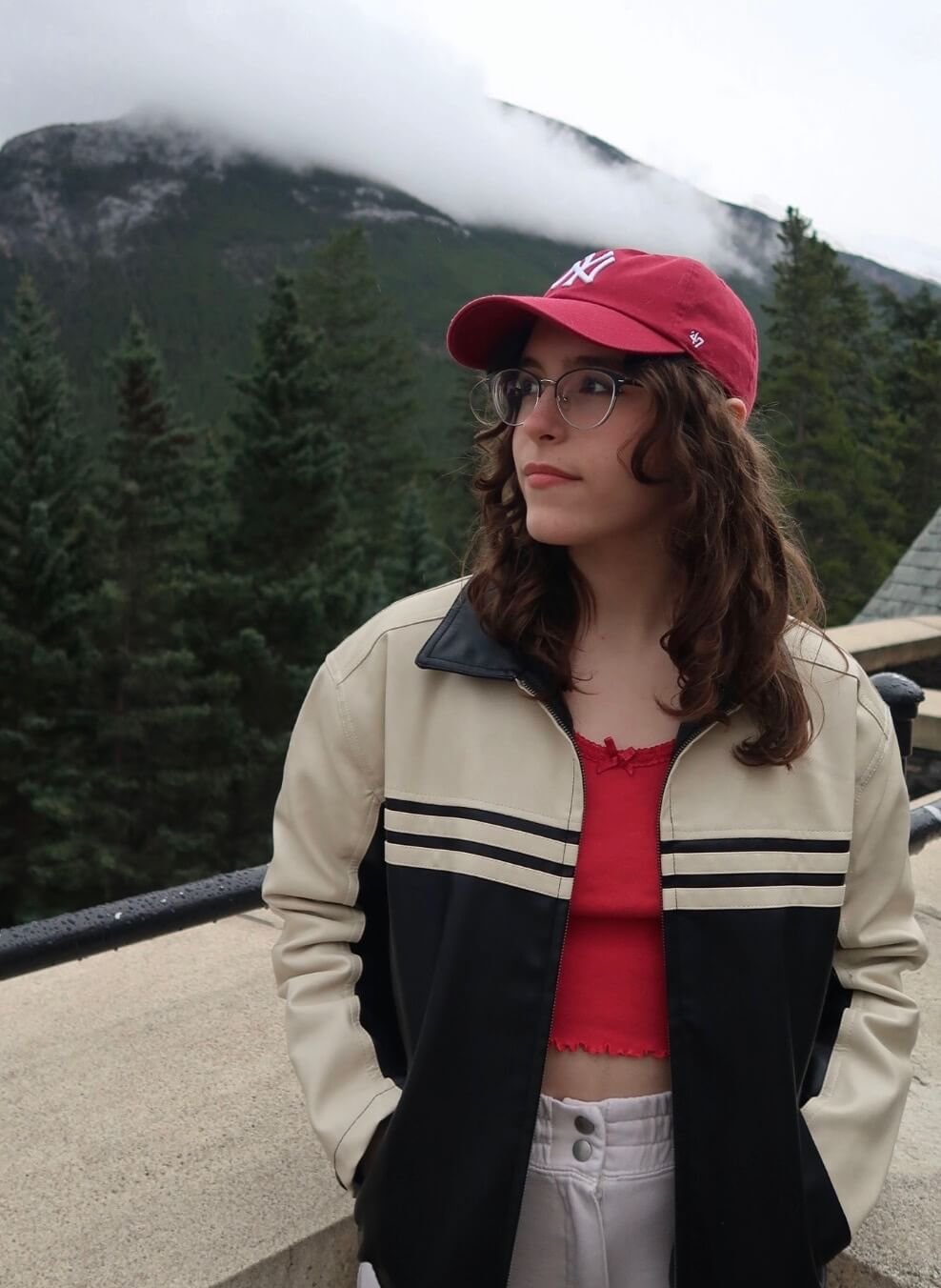 Young woman with curly brown hair, glasses, red cap, beige and black jacket, red crop top, and white pants standing outdoors near a fence with a forest and mountain with clouds in the background.