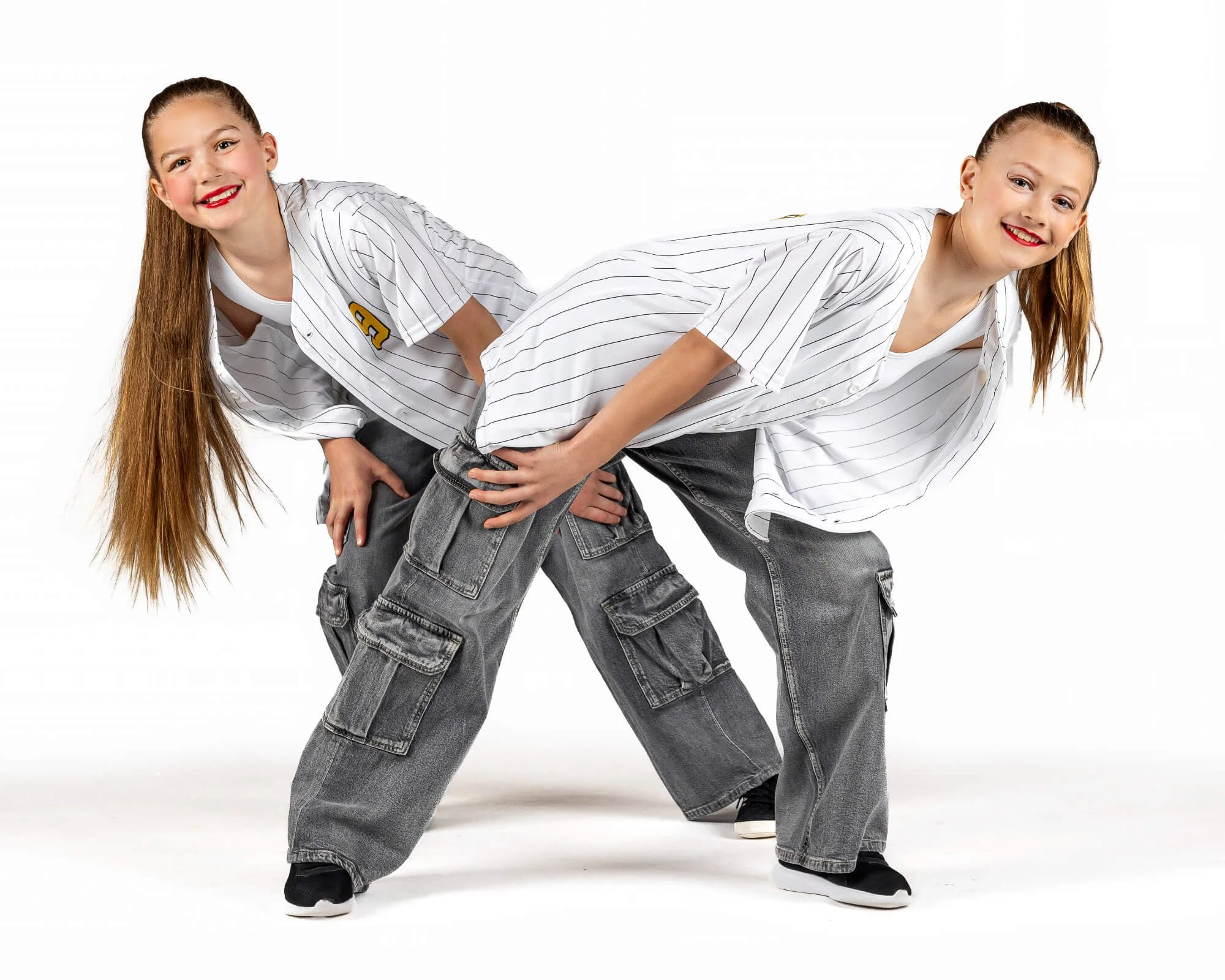 Two young girls engaging in a playful wrestling pose, both wearing white baseball jerseys with pinstripes and gray cargo pants, on a white background.