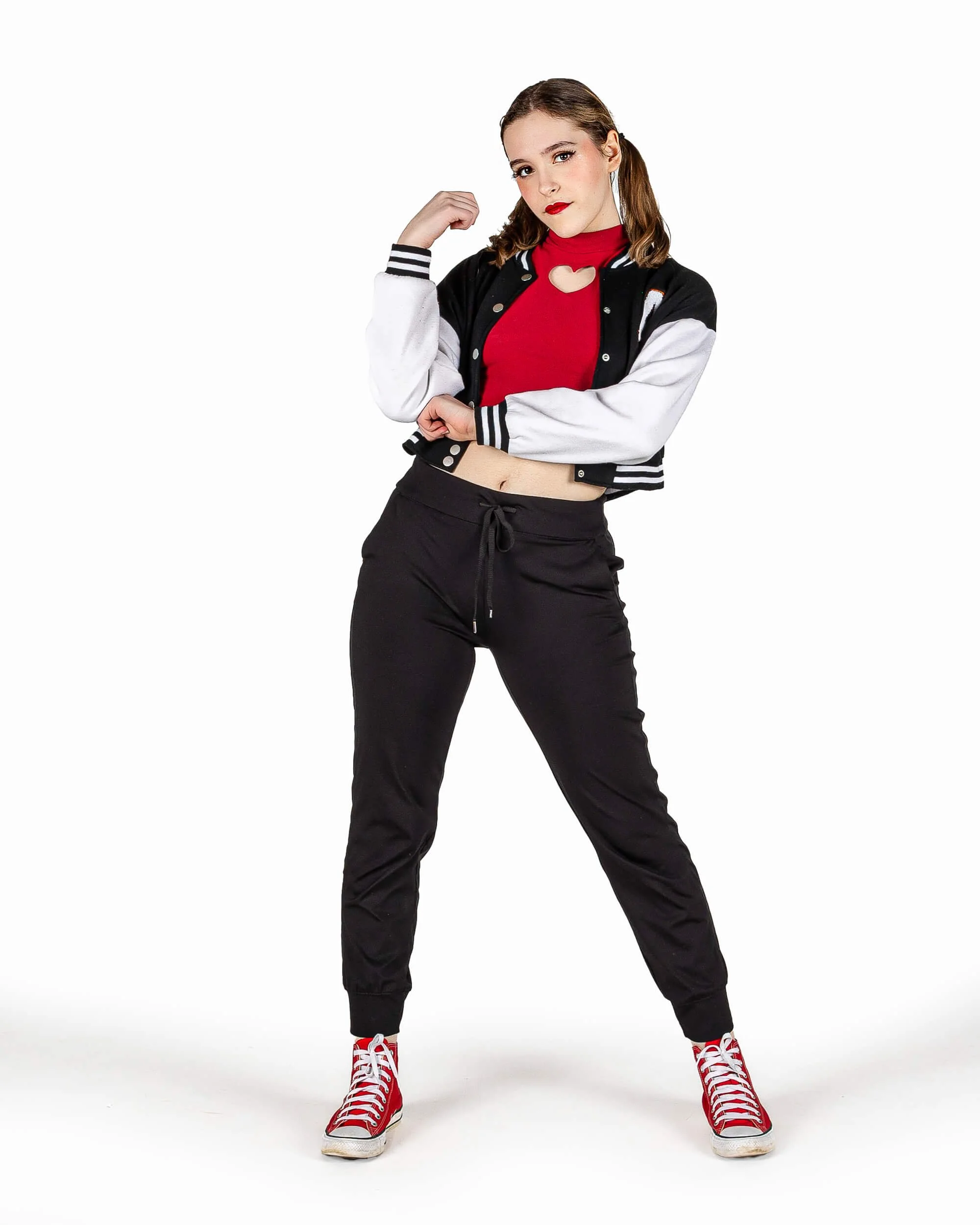 A young woman with light brown hair in pigtails, wearing a black and white bomber jacket, red crop top with a heart-shaped cutout, black jogger pants, and red sneakers, standing with one arm flexed and the other crossed, looking confidently at the camera.