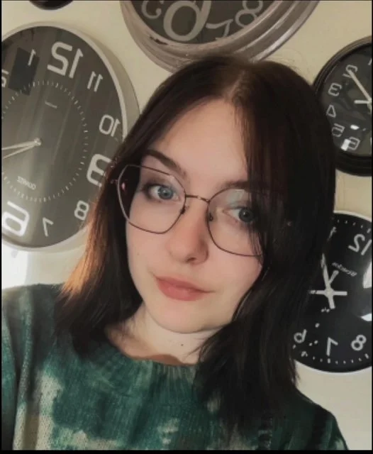 Young woman with dark hair and glasses taking a selfie in front of multiple wall clocks.