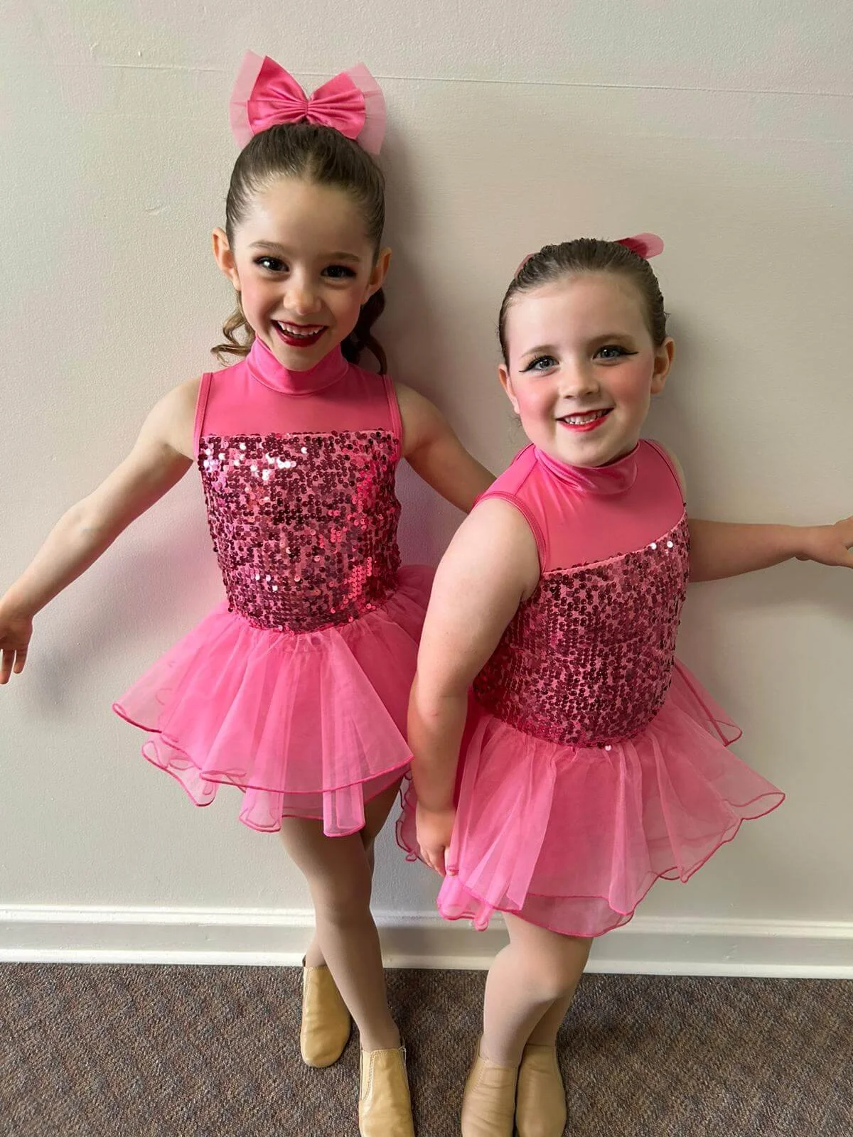 Two young girls dressed in pink dance costumes with sequin tops, tulle skirts, and tan dance shoes, standing against a plain wall. Both girls have their hair tied back with large pink bows and are smiling at the camera.