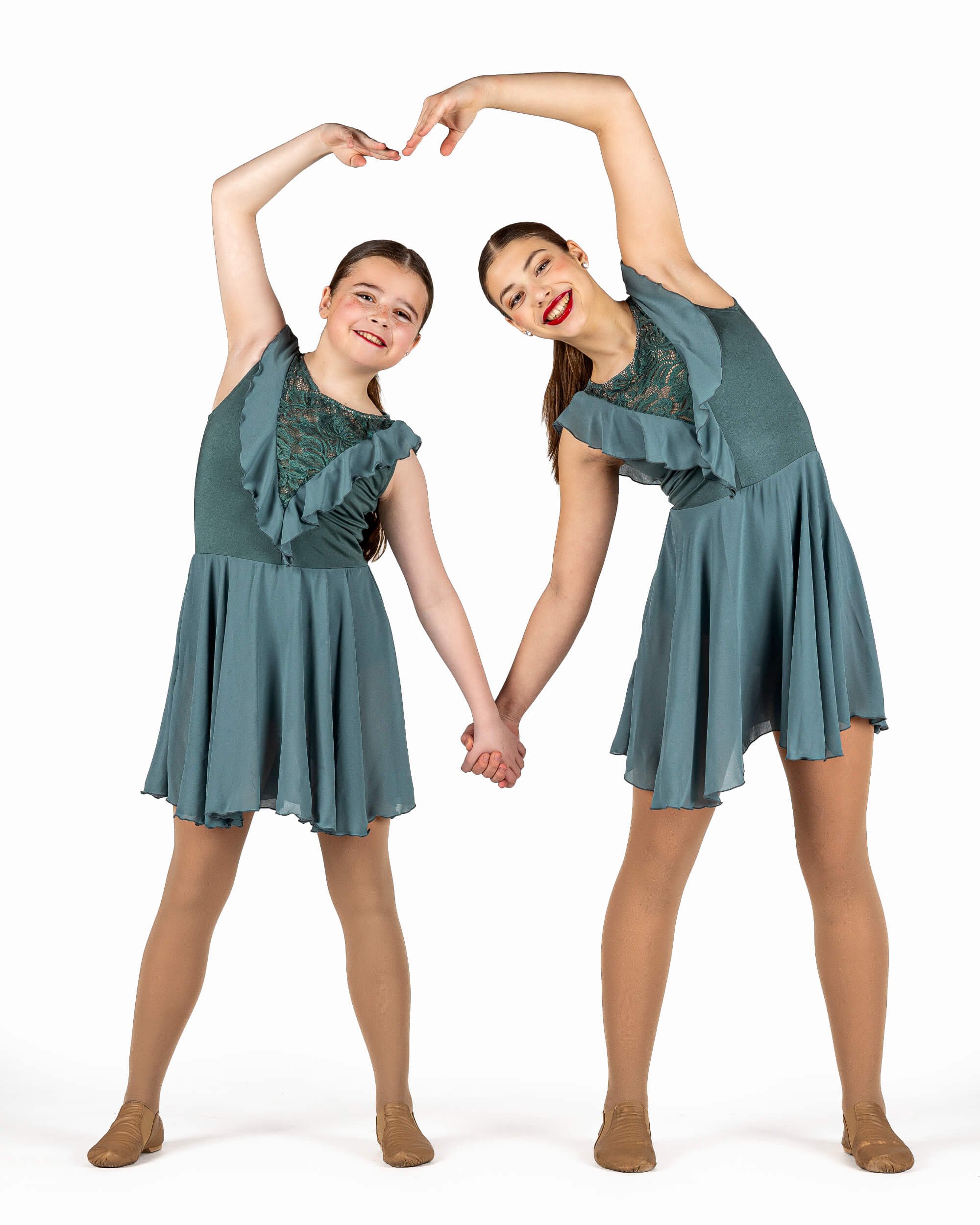 Two young girls in matching teal dance costumes holding hands and forming a heart shape with their arms, smiling happily.