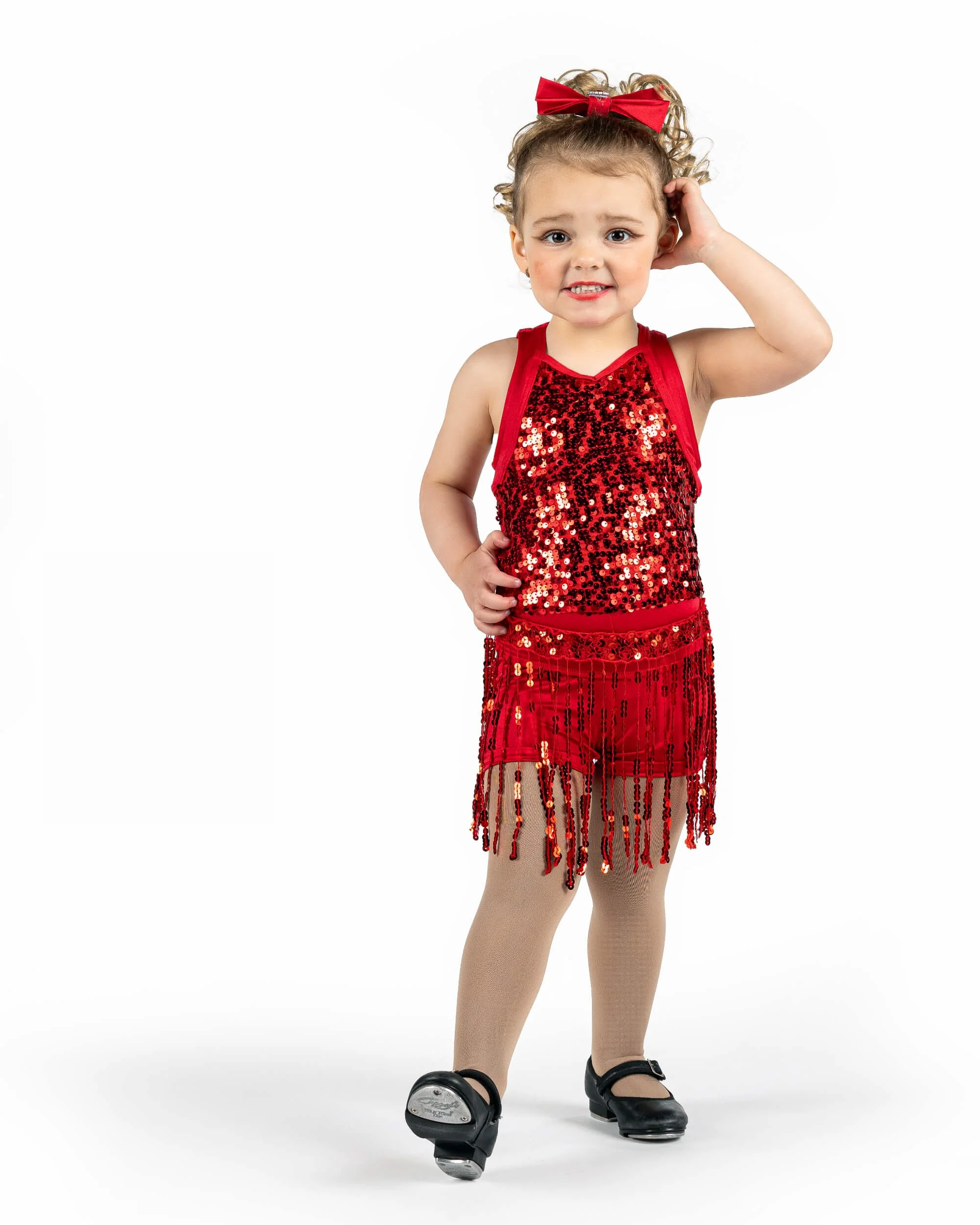 A young girl with curly blonde hair wearing a red sequin dress with fringe, tan tights, black tap shoes, and a red bow in her hair, standing against a white background for her dance portrait.