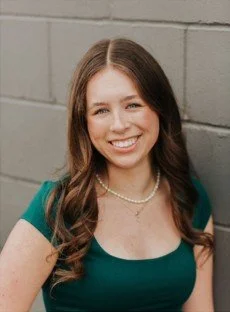 A young woman with long brown hair smiling and standing against a gray brick wall wearing a teal dress and a pearl necklace.