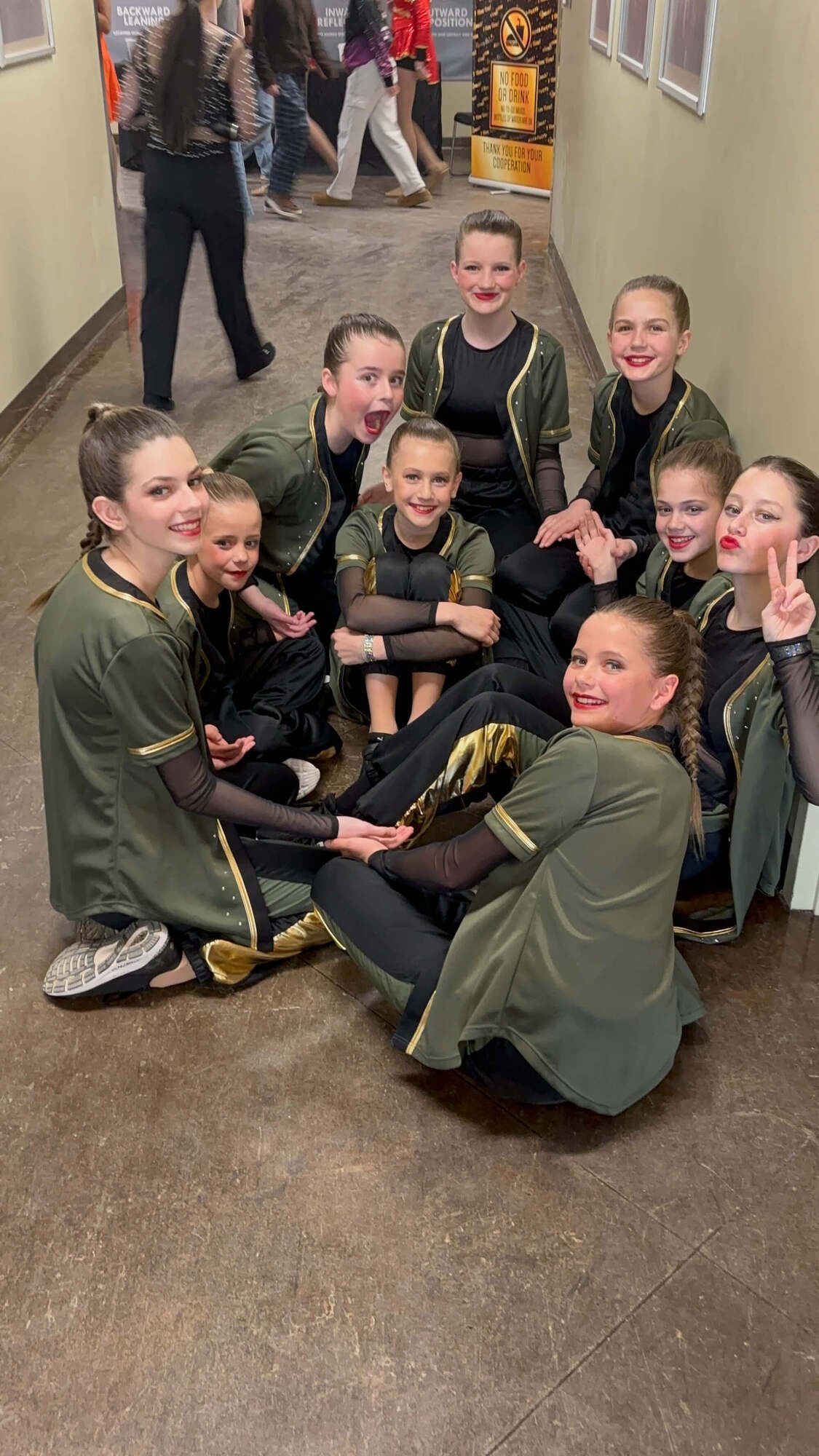 A group of young girls in matching dance or cheer uniforms sitting on the floor in a circle, holding hands, posing happily for a photo.