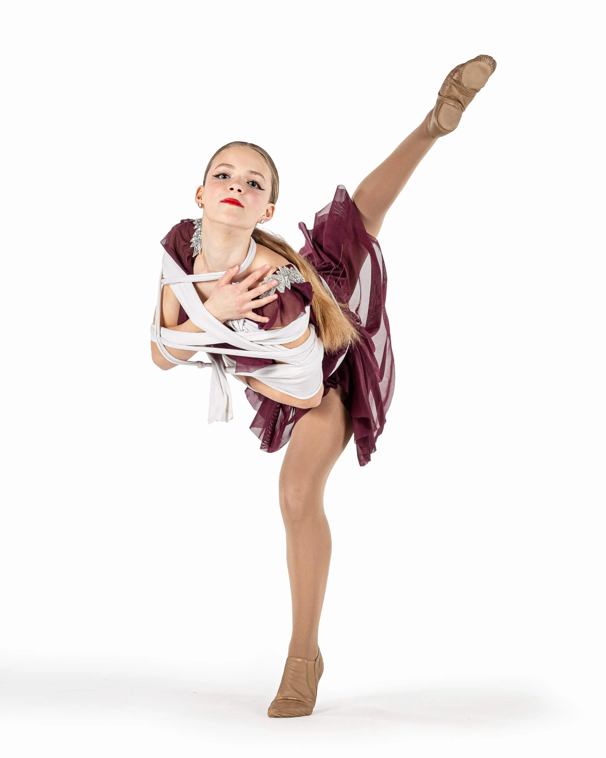 Young girl performing a ballet dance pose in a maroon and white costume with beige ballet shoes, standing on one leg with the other leg extended high, against a white background.