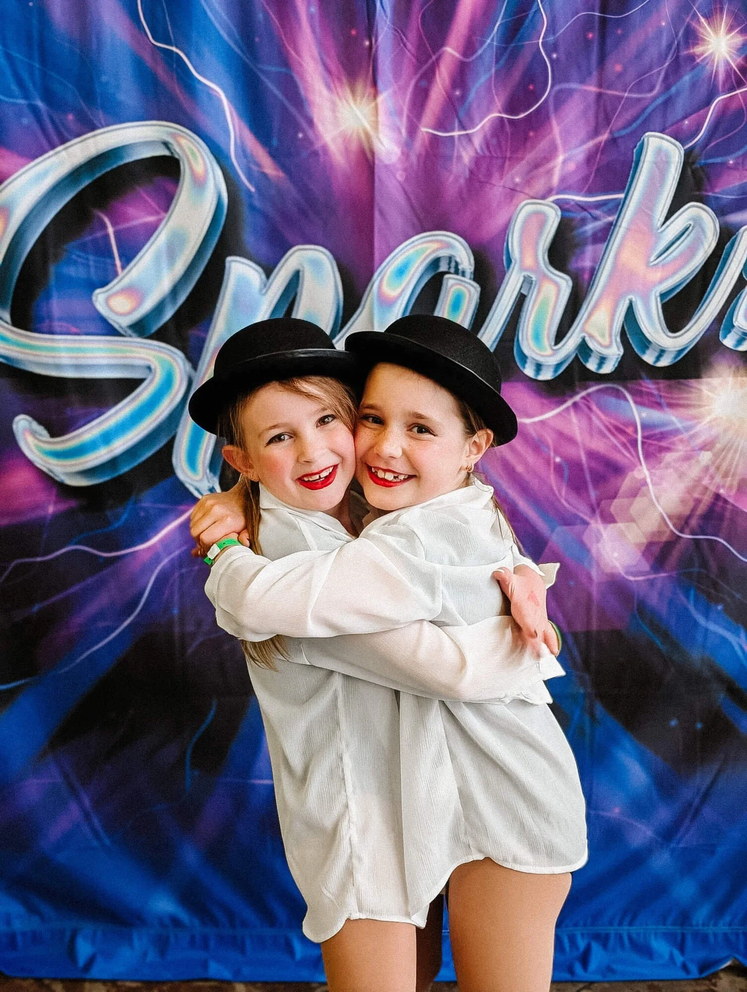 Two young girls hugging each other in front of a colorful backdrop that says "Dance" and "Spark." They are wearing white outfits and black hats, smiling and showing their teeth.