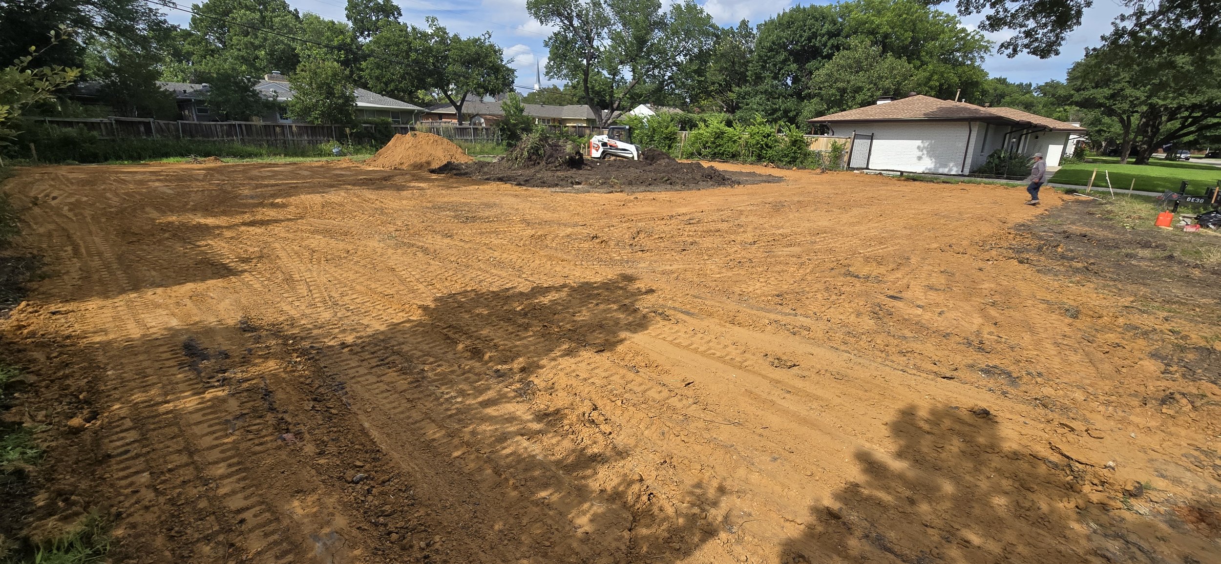 A cleared construction site with orange dirt and tire tracks, a small pile of dirt, and construction workers in the background. Trees and residential houses surround the site.