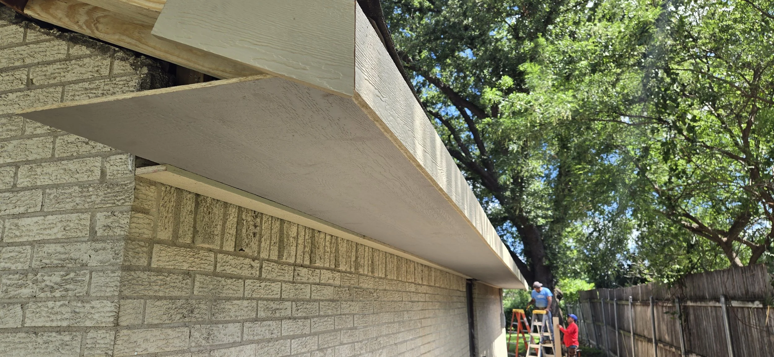 Close-up view of a newly installed white soffit under a roof overhang, with brick exterior wall and a backyard scene with workers and trees visible in the background.