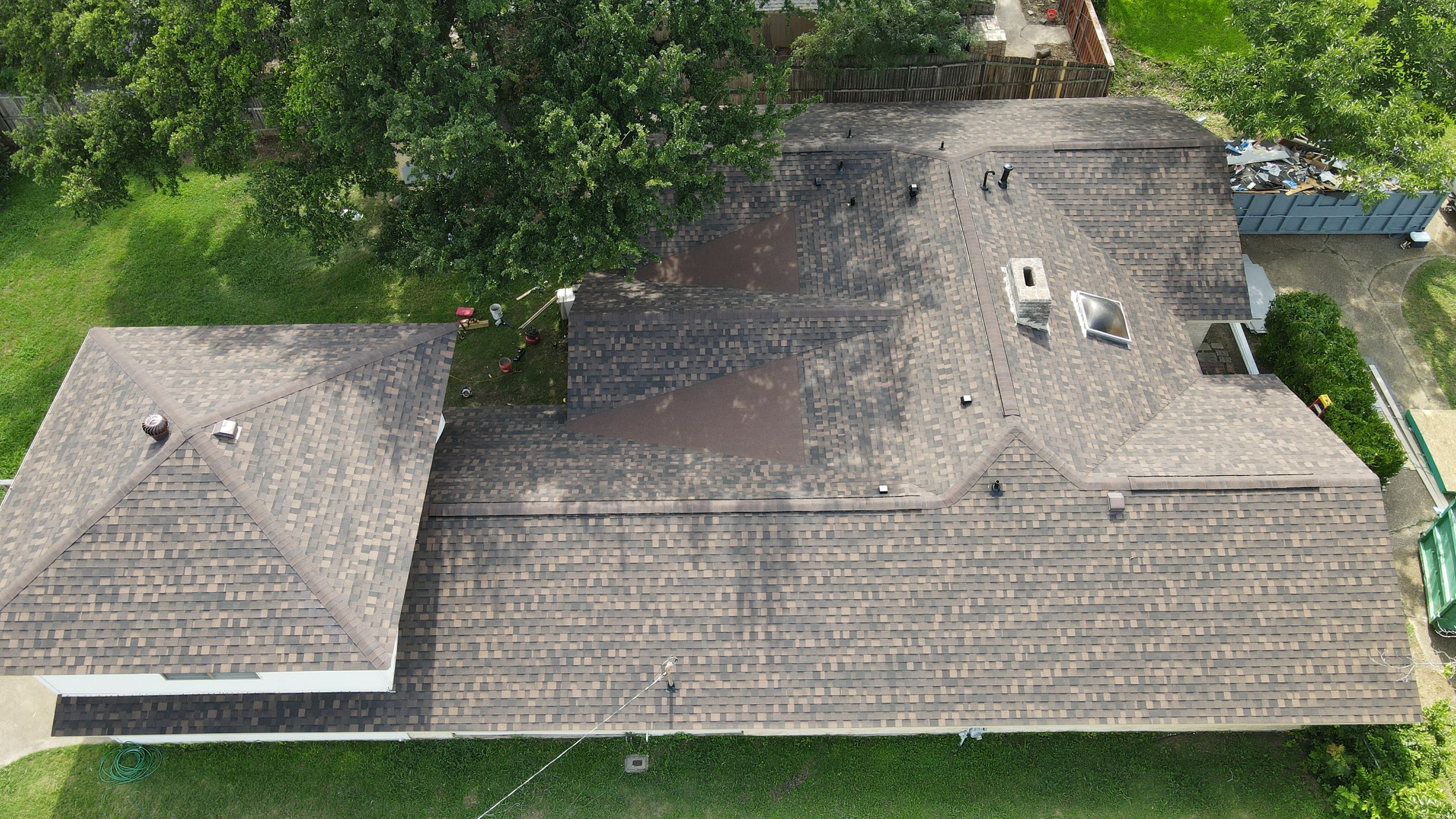 An aerial view of a house with a shingled roof, surrounded by green trees and grass, with parts of the yard and neighboring yards visible.