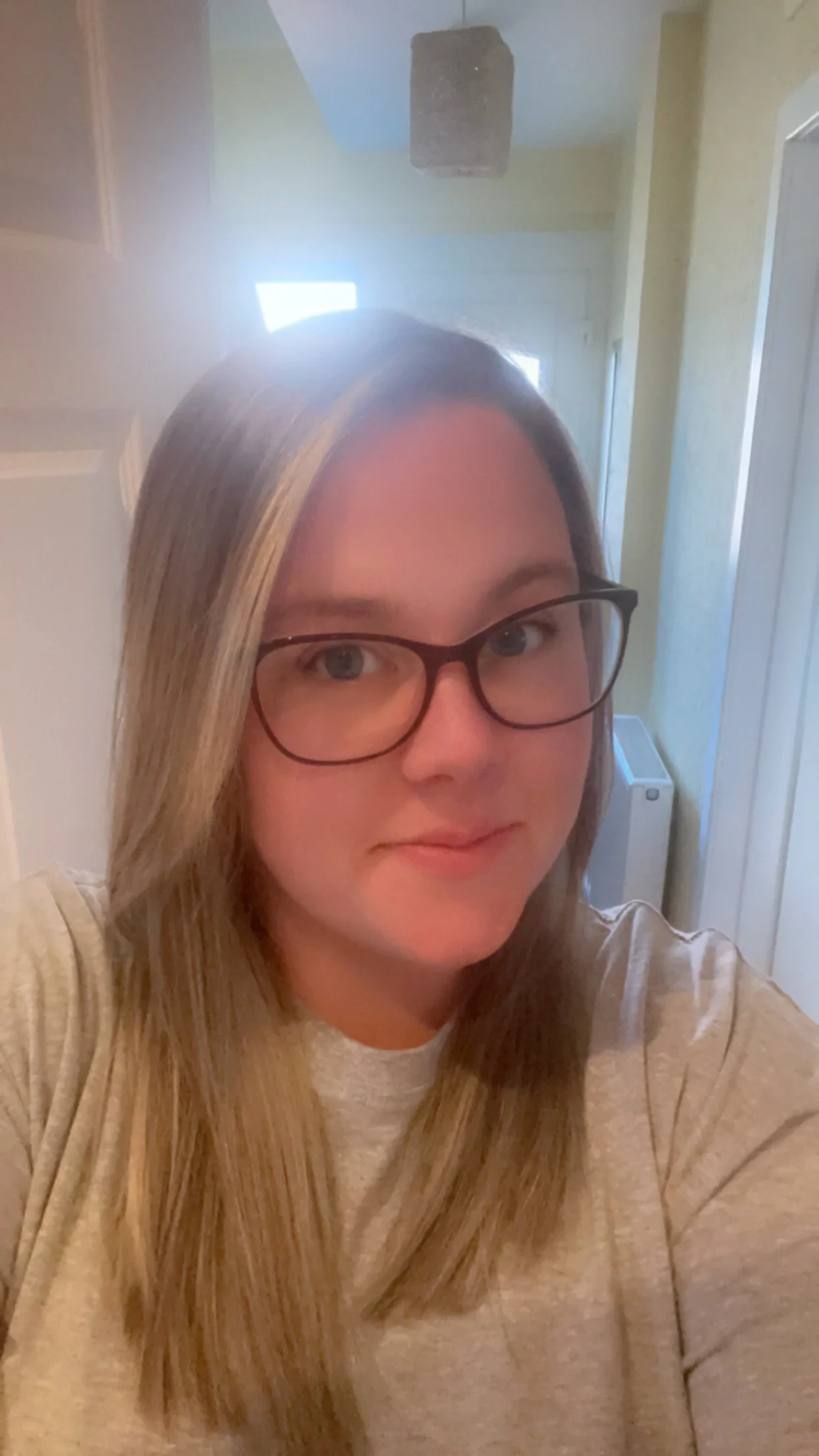 A woman wearing glasses with long, straight blonde hair, smiling gently in a room with a door, a window, and a heater visible in the background.