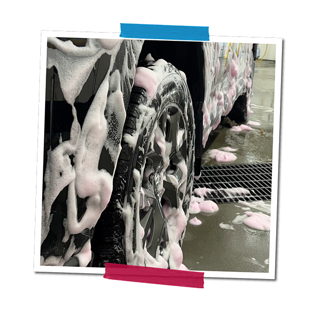 Close-up of a car wheel covered in soap suds during a car wash, with soap bubbles and foam on the ground.