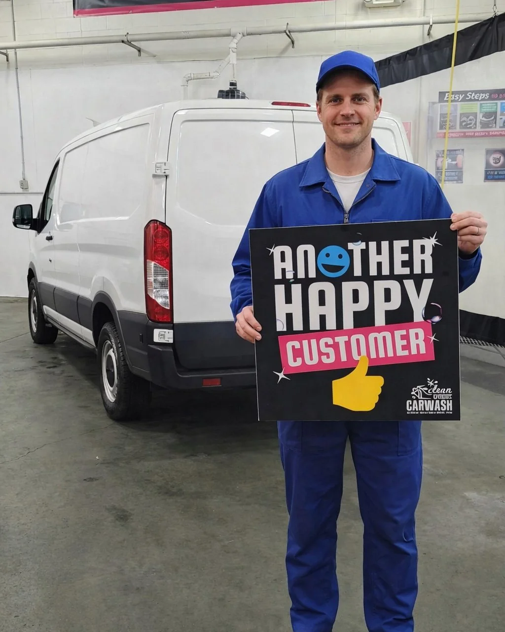 A smiling man dressed in a blue uniform and cap standing in a garage, holding a sign that reads "Another Happy Customer" with a yellow thumbs-up graphic and a logo for a car wash.