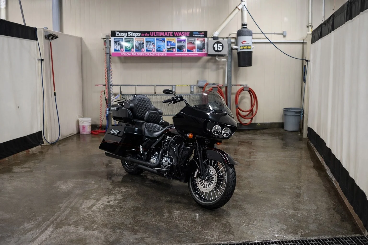 A black touring motorcycle with a large front fairing and comfortable seating is parked inside a car wash bay with concrete floor and white and black interior walls. There are hoses on the wall, a bucket, a trash can, and signs about washing instructions.