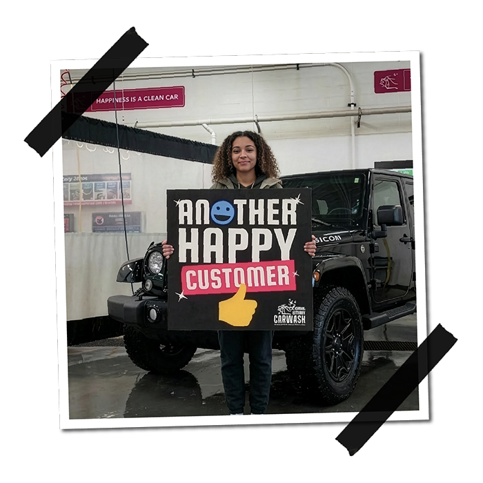 A woman standing in front of a black Jeep at a car wash while holding a sign that reads 'Another Happy Customer'. The background shows a car wash interior with a white wall and a purple sign that says 'Happiness is a clean car'.