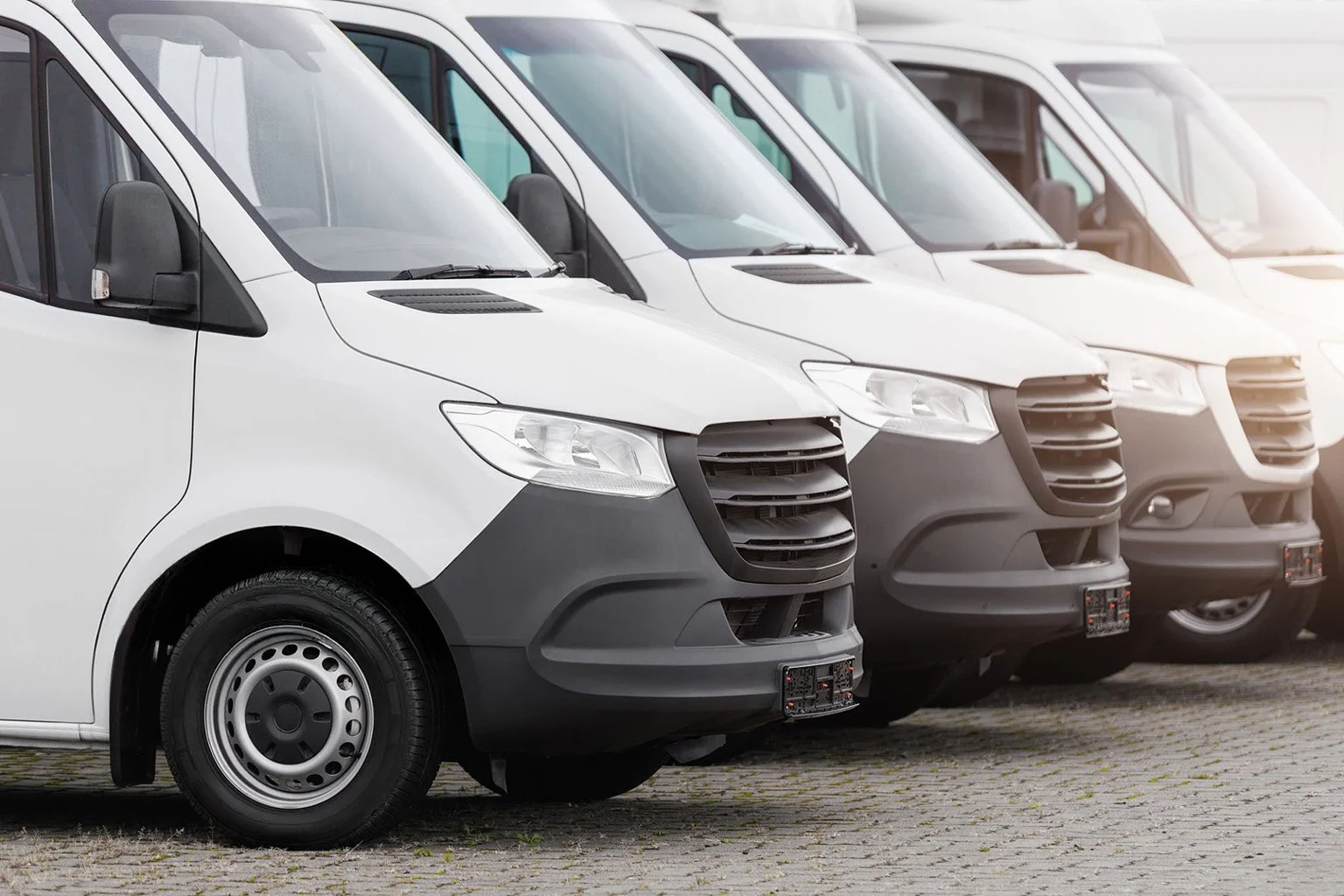 Several white commercial vans parked in a row on a cobblestone surface.