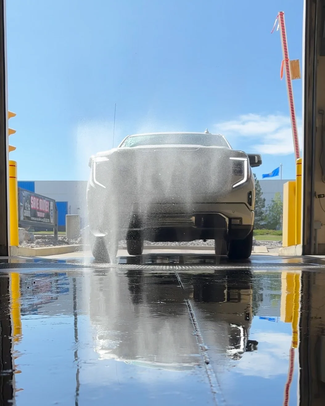 Car going through an automatic car wash with water spray, blue sky, and flags in the background.