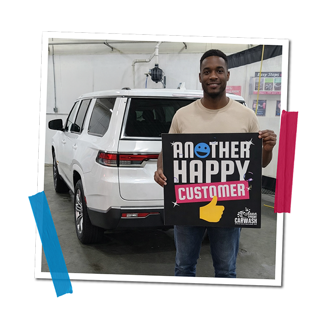 A smiling man holding a sign that says 'Another Happy Customer' with a thumbs-up icon inside a car wash facility.