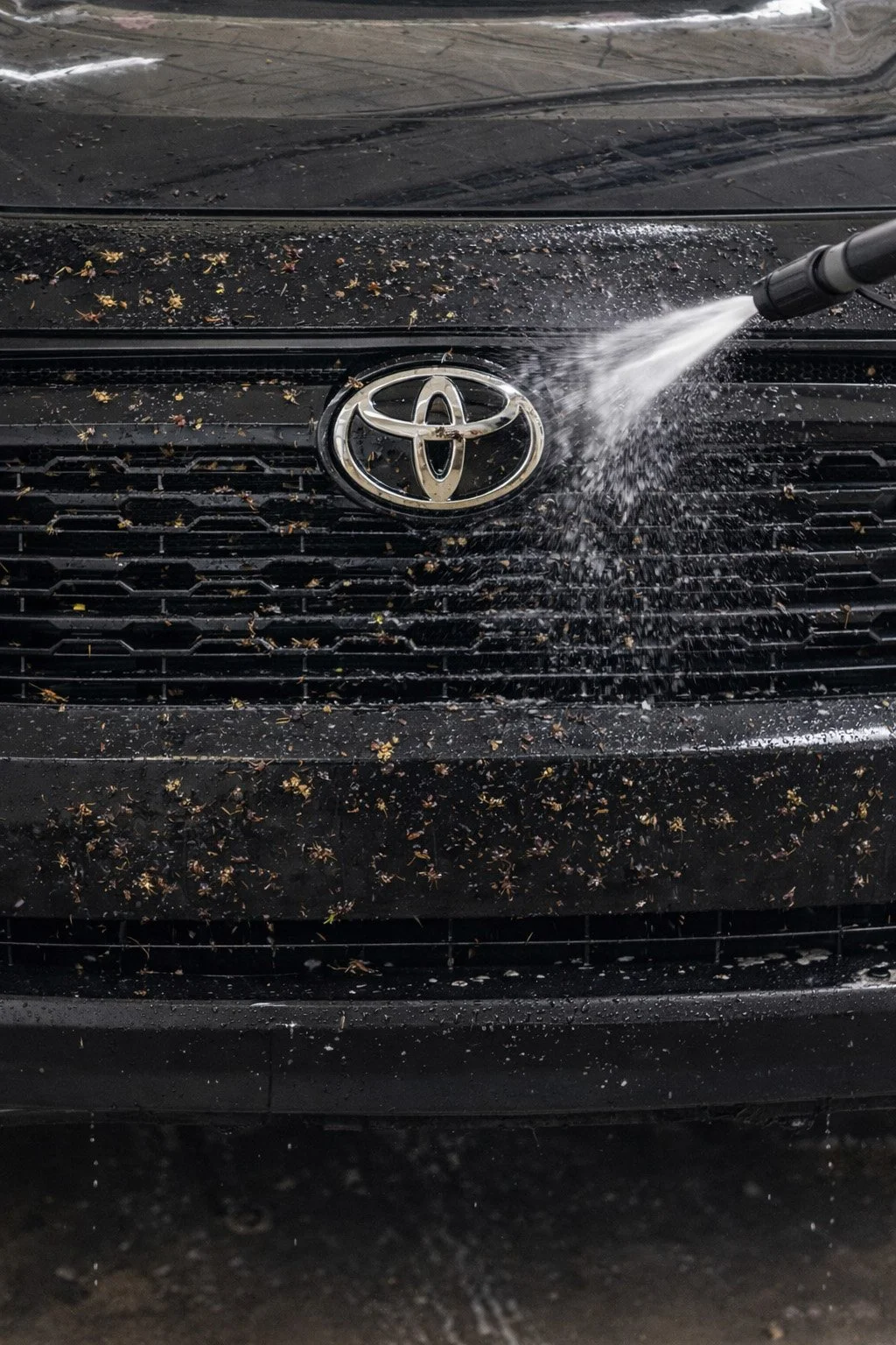 A black Toyota car being washed with water from a high-pressure spray, with water and dirt on the front grill and hood.