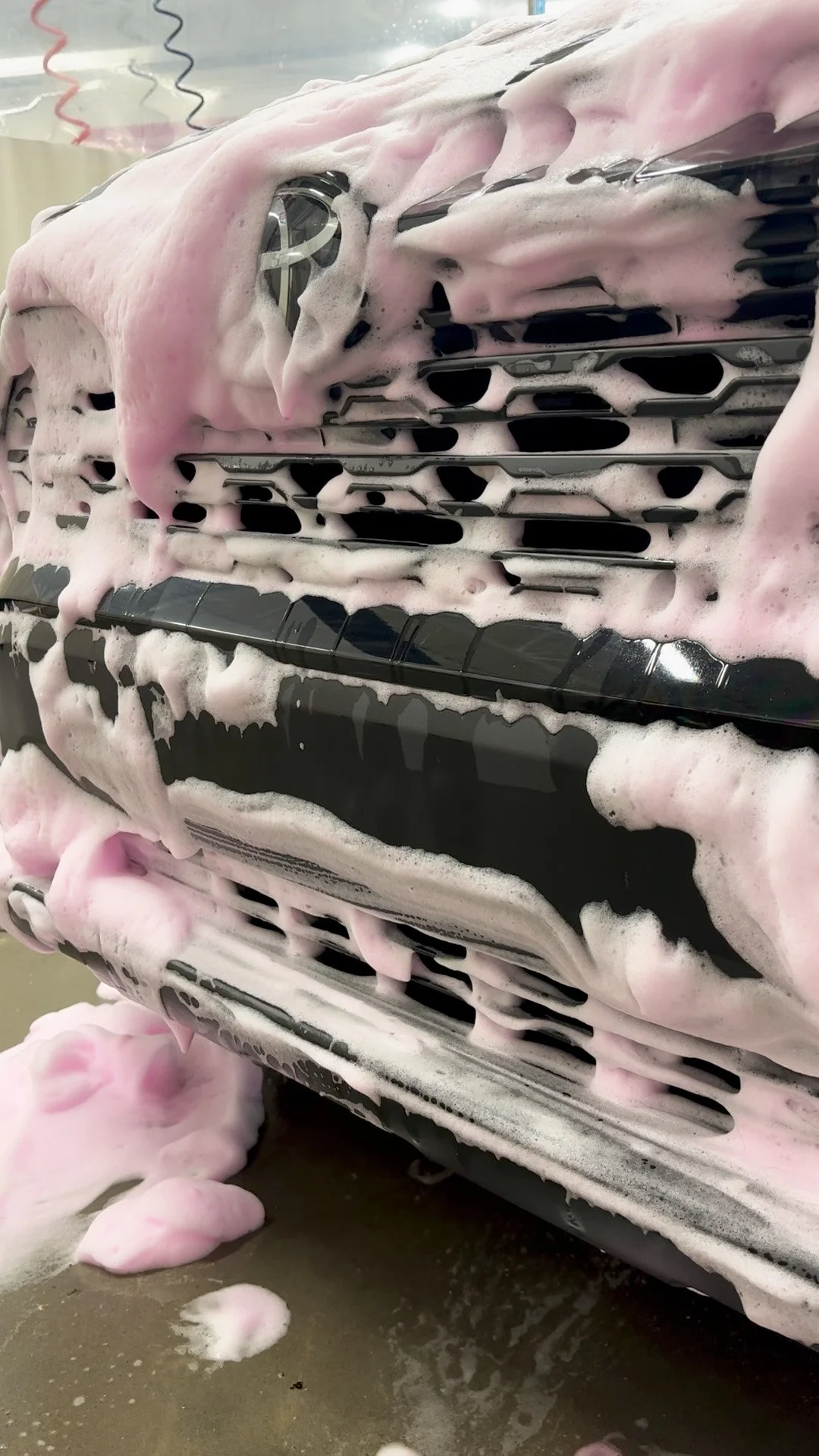Close-up of a black car being washed with pink foam and soap suds covering the front grille.