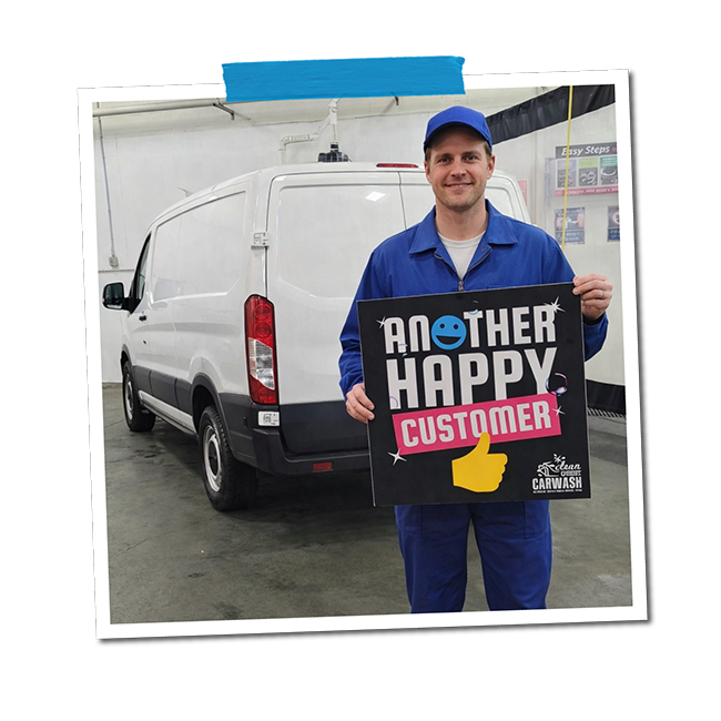 A man in blue coveralls and a blue cap holding a sign that reads 'Another Happy Customer' with a smiling face icon and a thumbs-up symbol, standing inside a car wash facility next to a white van.