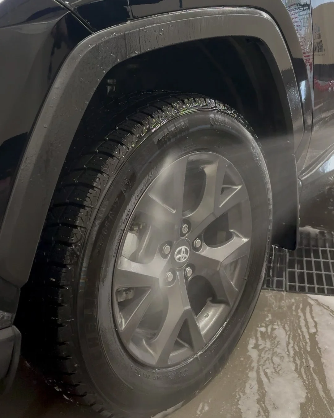Close-up of a black car's tire and rim being washed with steam, showing water droplets and a wet floor.
